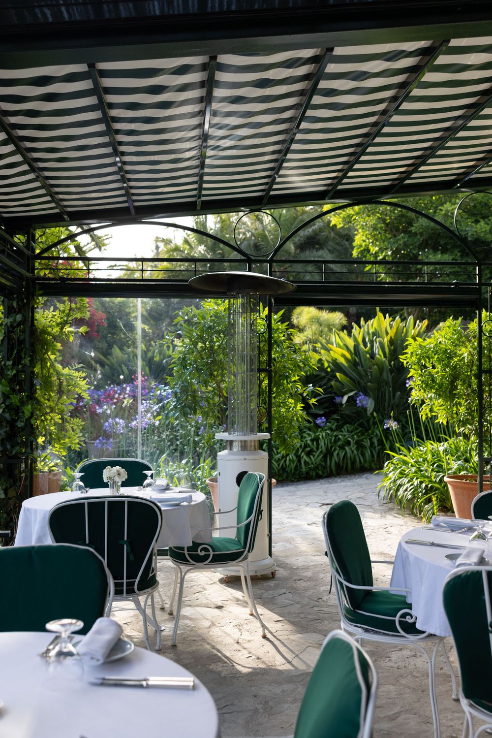 Garden restaurant terrace with green chairs and white tablecloths under striped canopy.