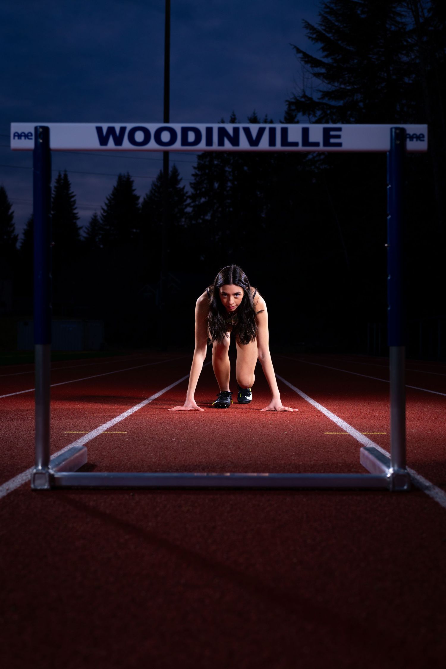 Athlete in starting position on running track beneath Woodinville sign during dusk with illuminated red surface.