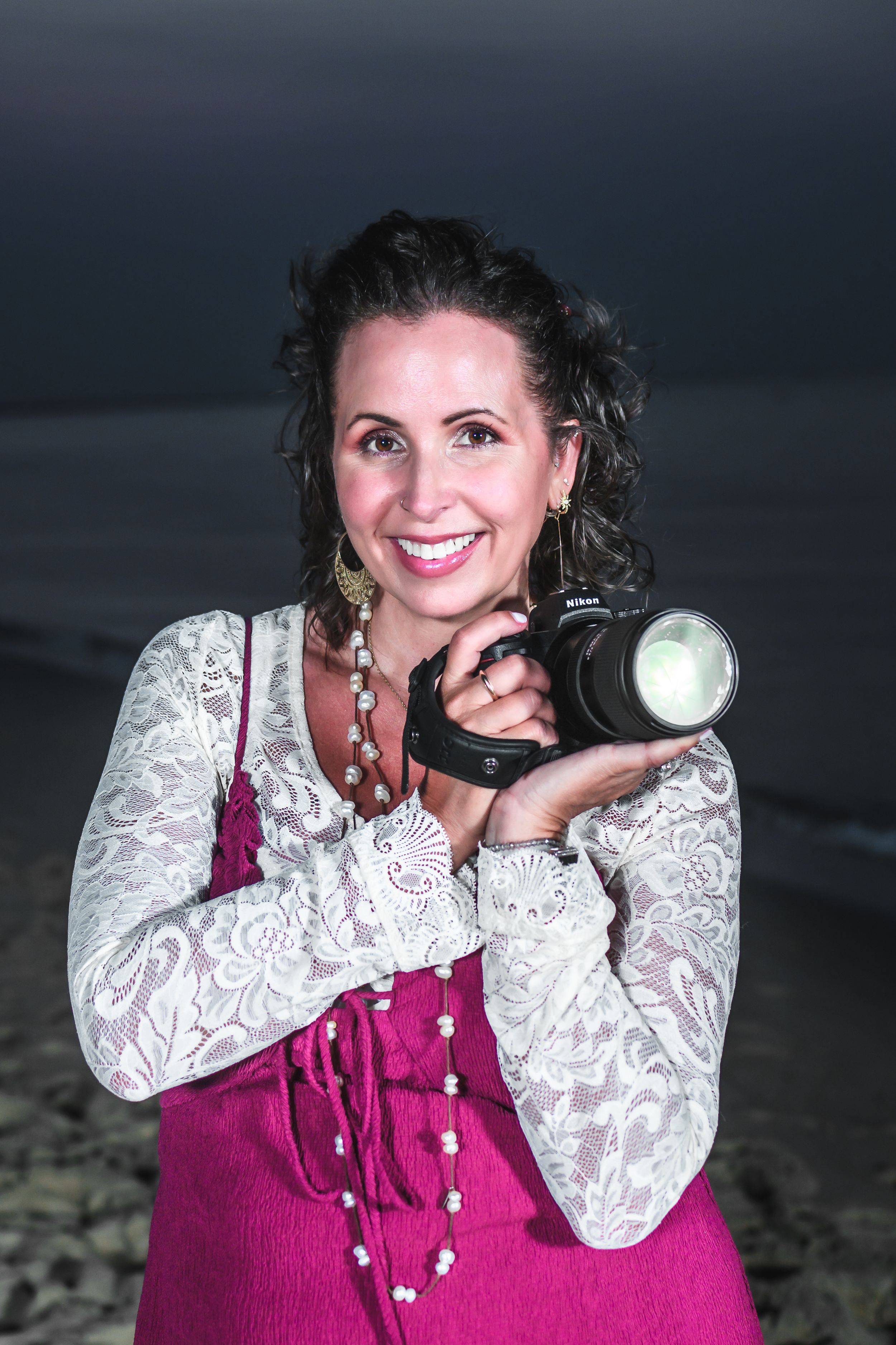 Photographer in white lace top and pink dress holding camera outdoors at night.