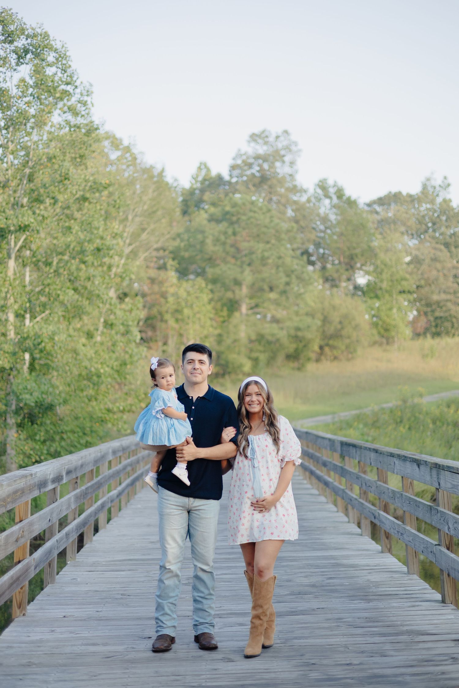 A couple stands together outdoors with their infant under a large tree.