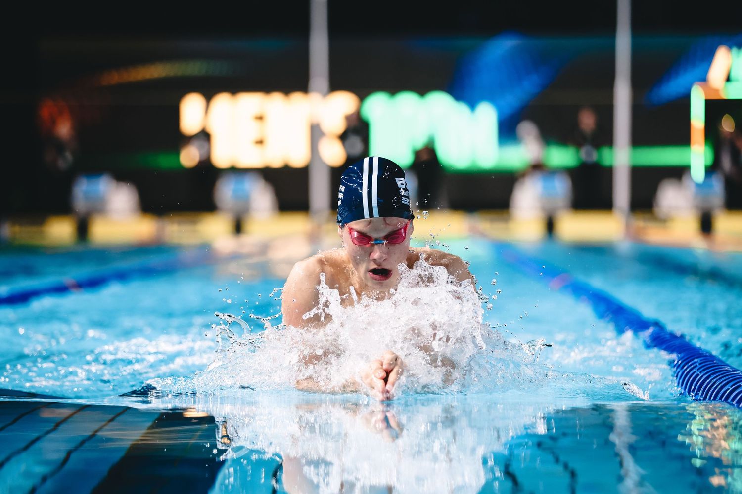 Swimmer performing breaststroke with dramatic water splash in nighttime pool setting.