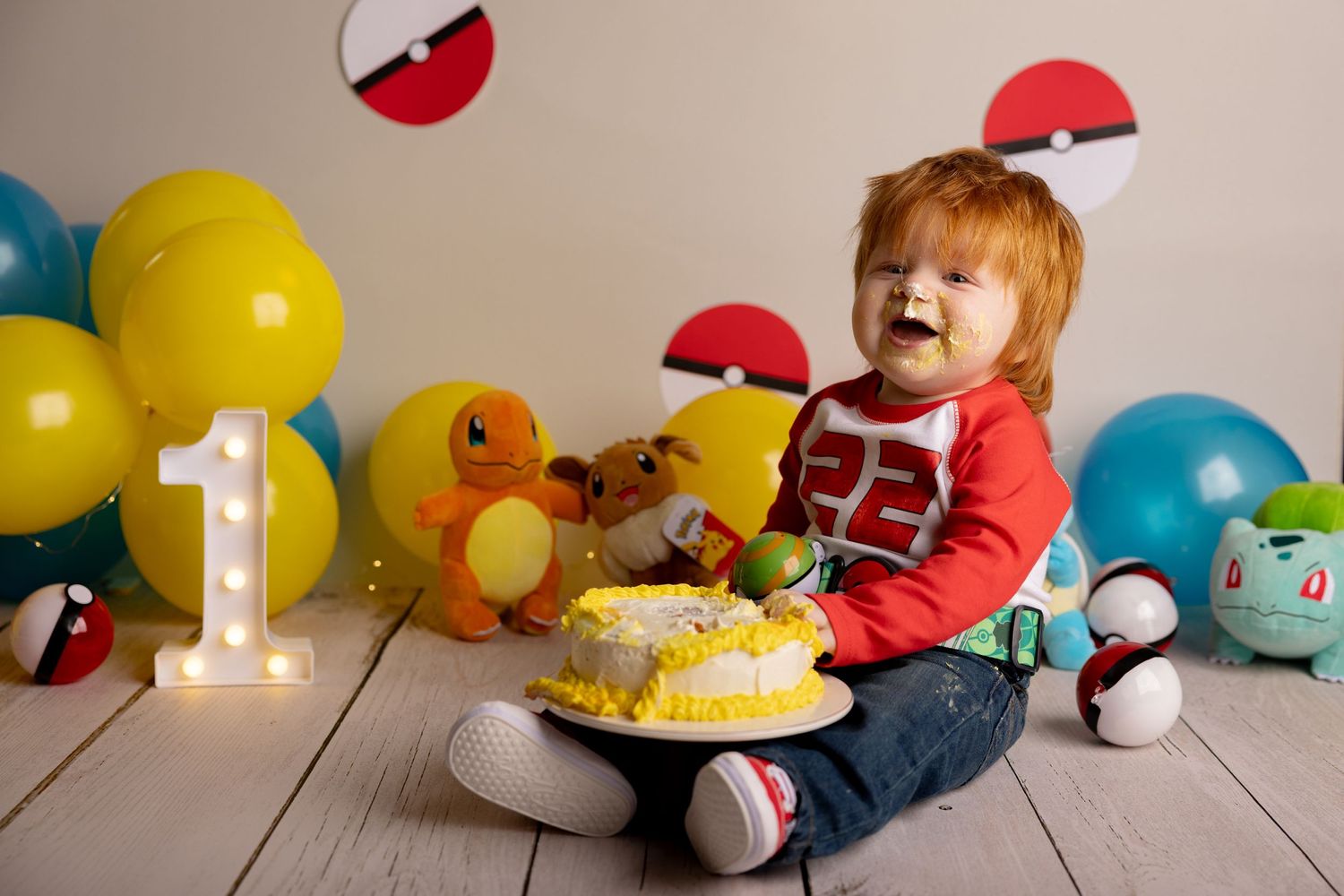 A first birthday party scene with colorful balloons, Pokemon plush toys, and a decorated cake on display.