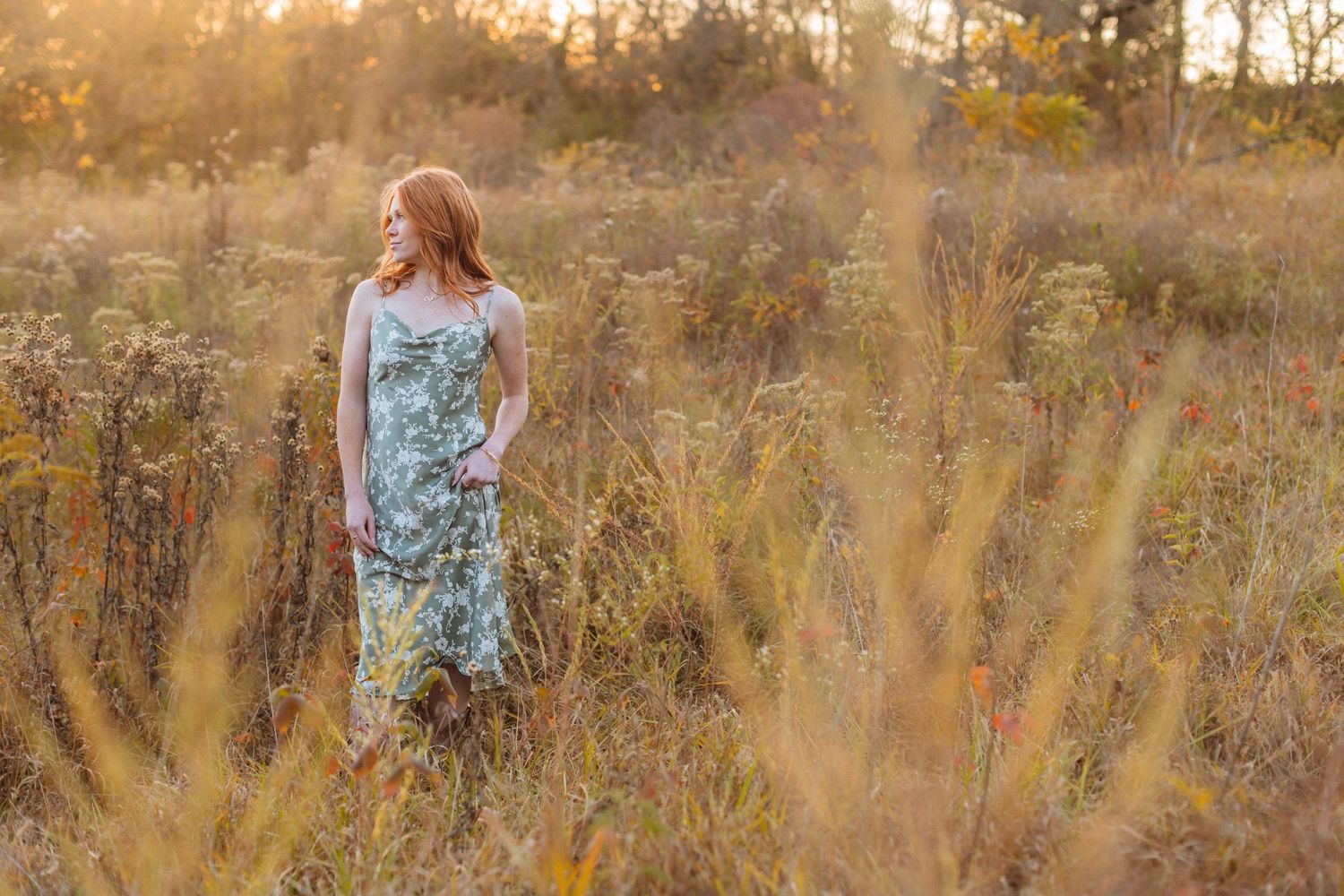 A young woman in floral green dress stands in a golden field of tall grasses and looks off to the side during a fall senior photo session.