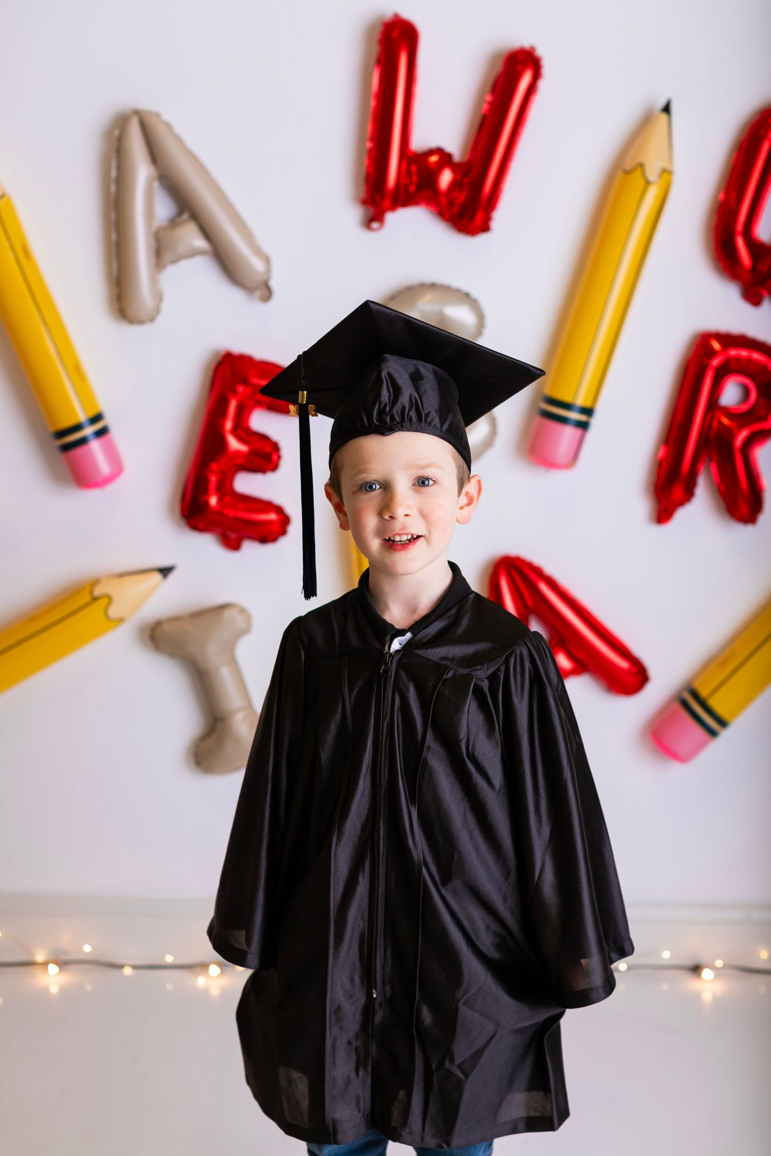 Student wearing graduation cap and gown poses against wall decorated with letters and pencil designs.