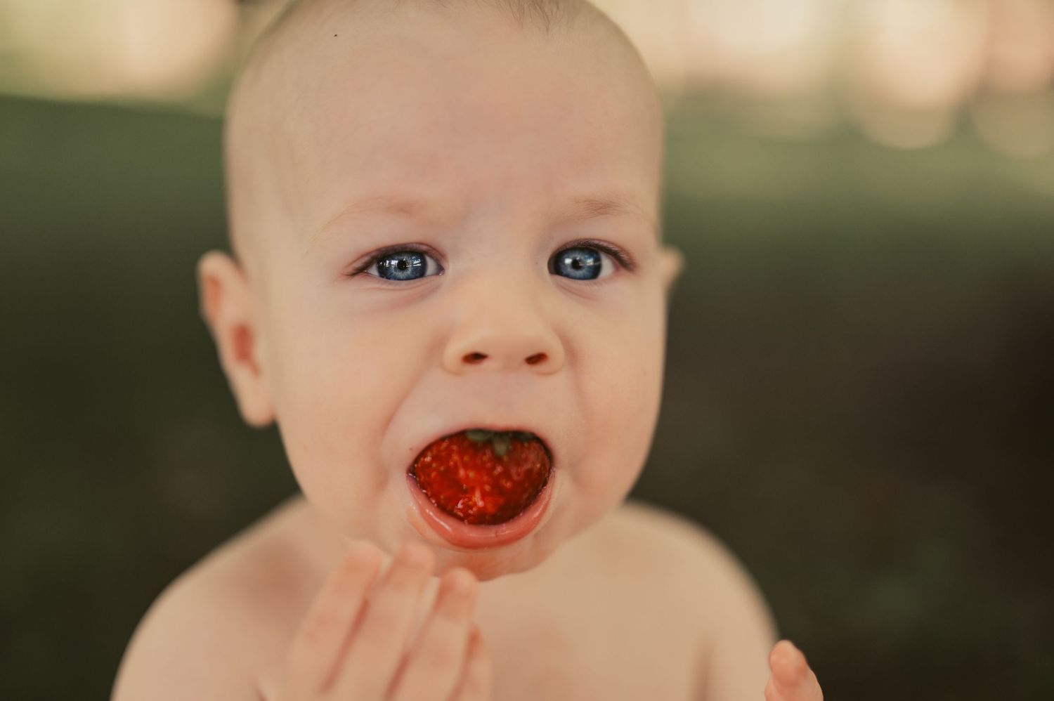 Blue-eyed baby eating a shiny red strawberry outdoors, juice on lips.