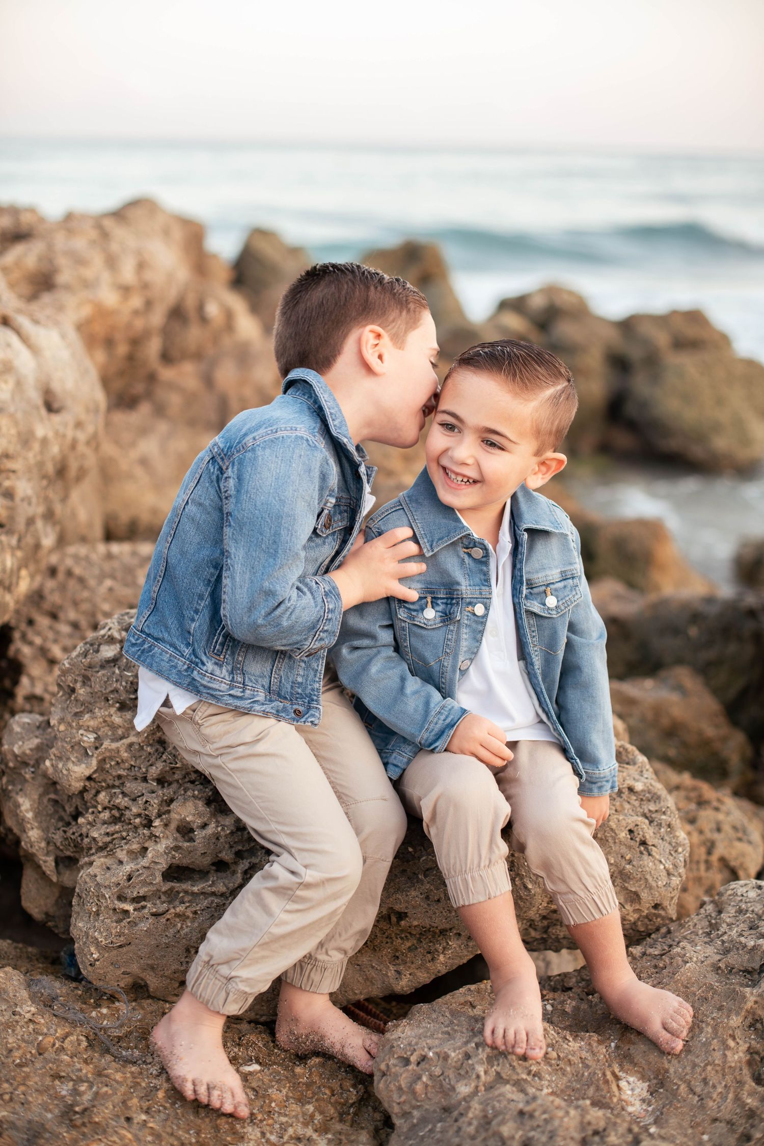 Two children in denim jackets share secrets while sitting on coastal rocks.