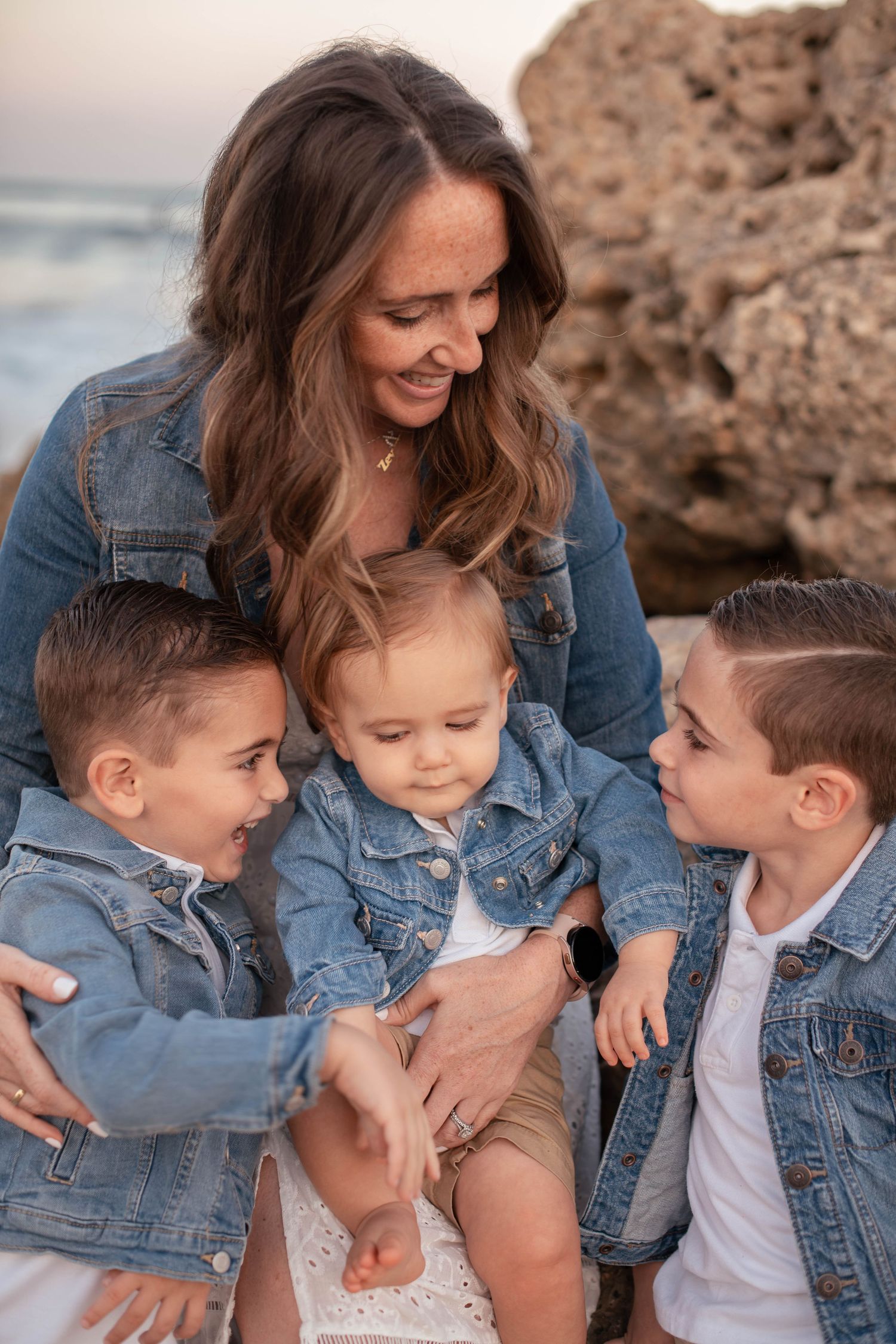 Family wearing matching denim jackets shares a tender moment by rocky beach at sunset.