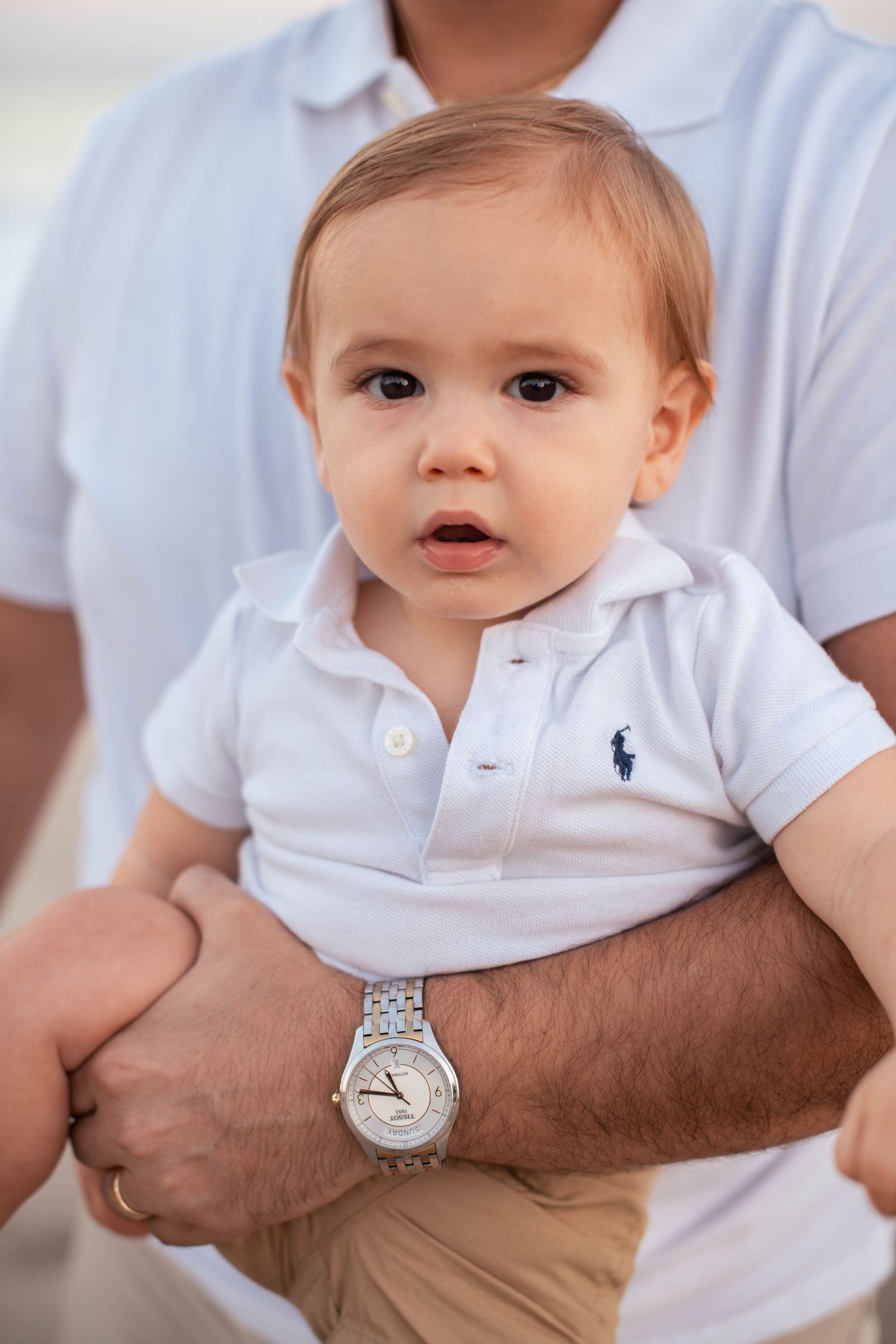 Baby in white polo shirt sits on lap wearing luxury watch.