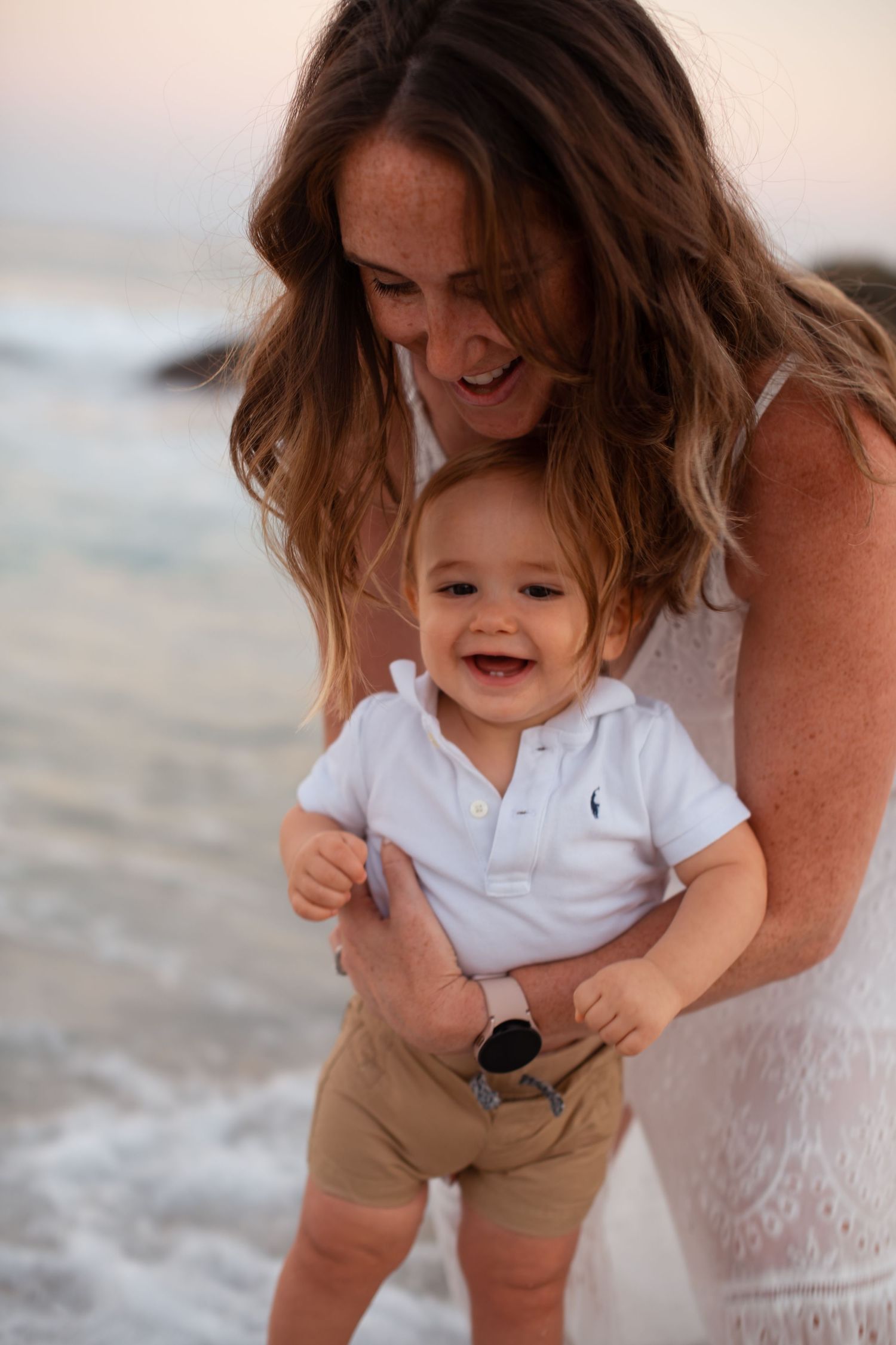 Mother and toddler share laughs by ocean waves during sunset photoshoot.