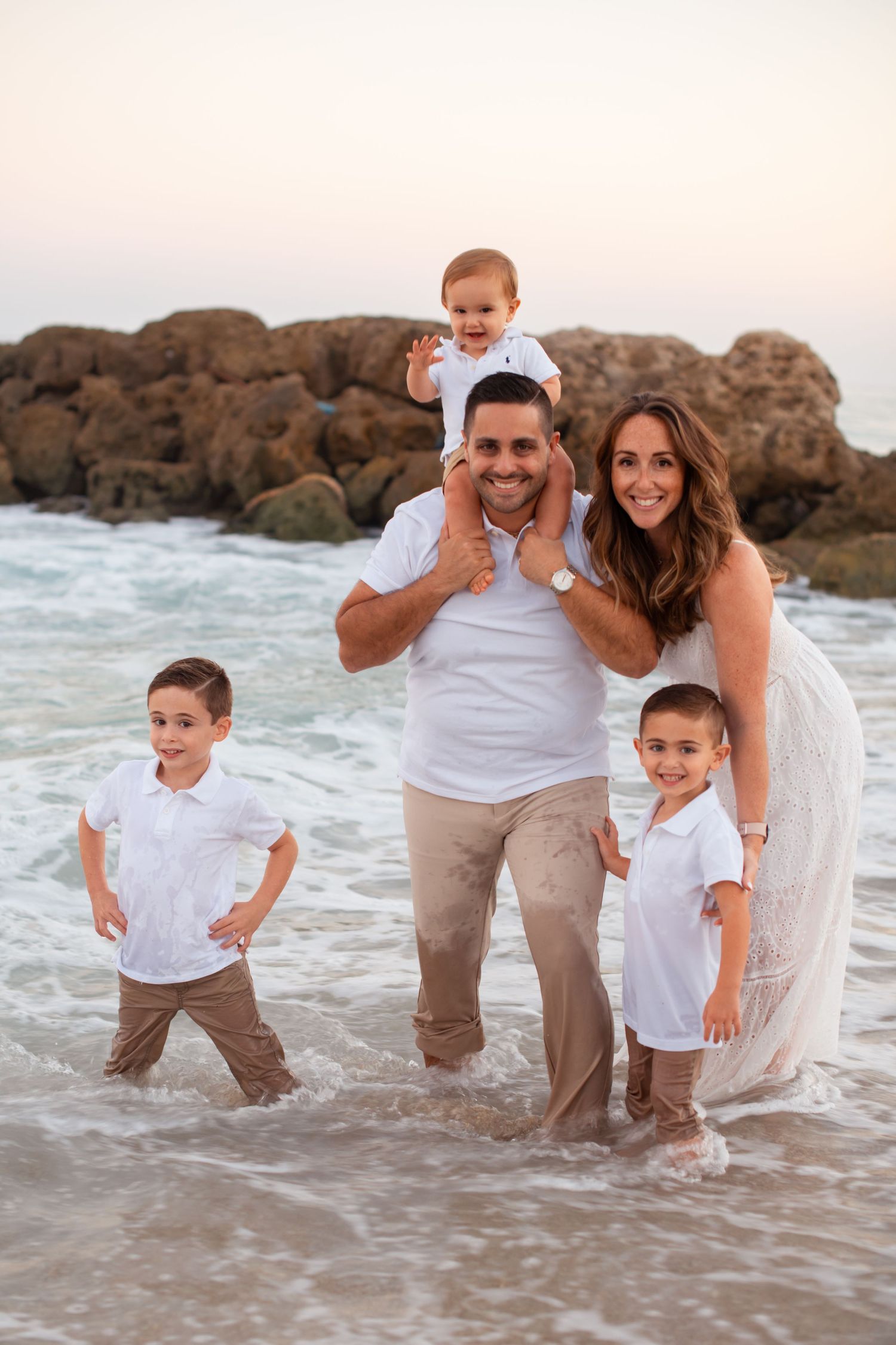Family of five stands together in ocean waves at sunset wearing coordinated white outfits.