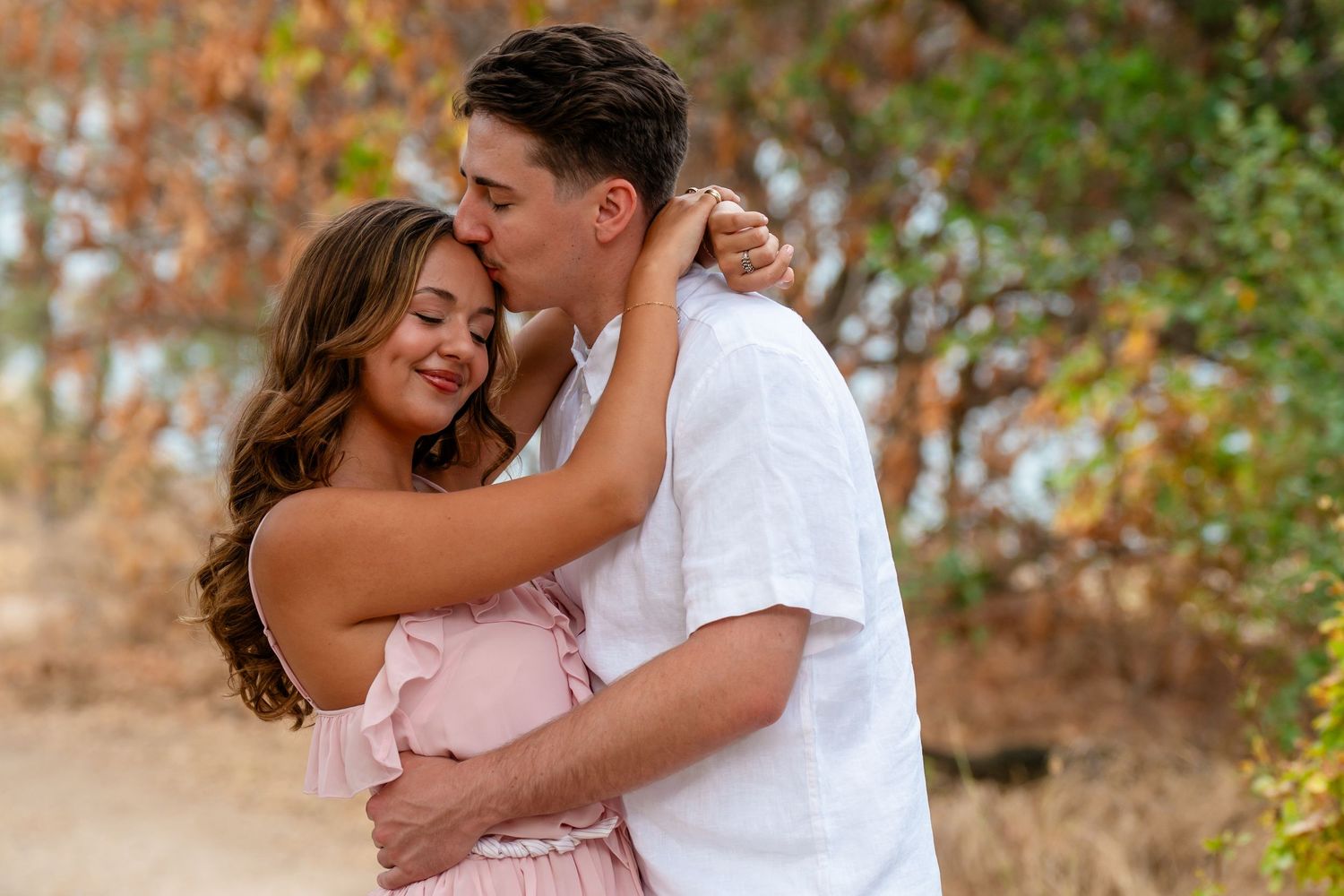 Romantic couple embracing outdoors, man kissing woman's forehead, wearing casual summer attire.