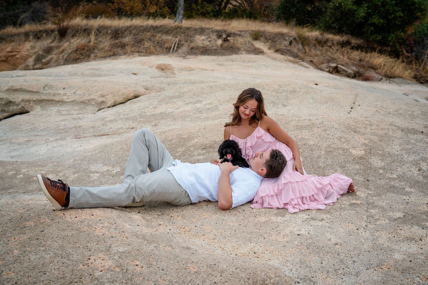 Couple relaxing on a large rock outdoors, woman in pink dress sitting beside reclining man in white pants.