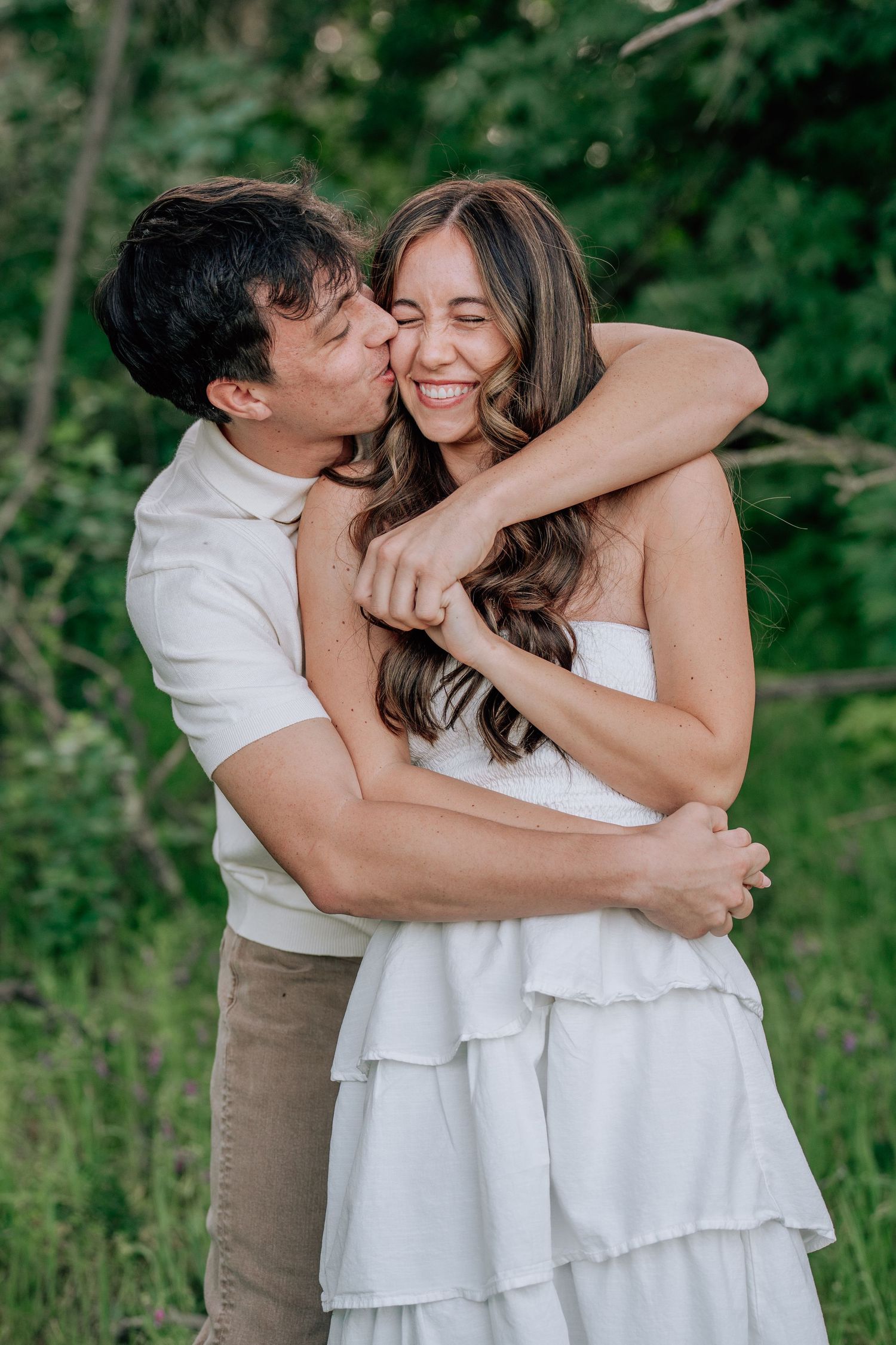 A couple embraces outdoors, man kissing woman's cheek as she smiles, wearing a white ruffled dress, with green foliage behind.