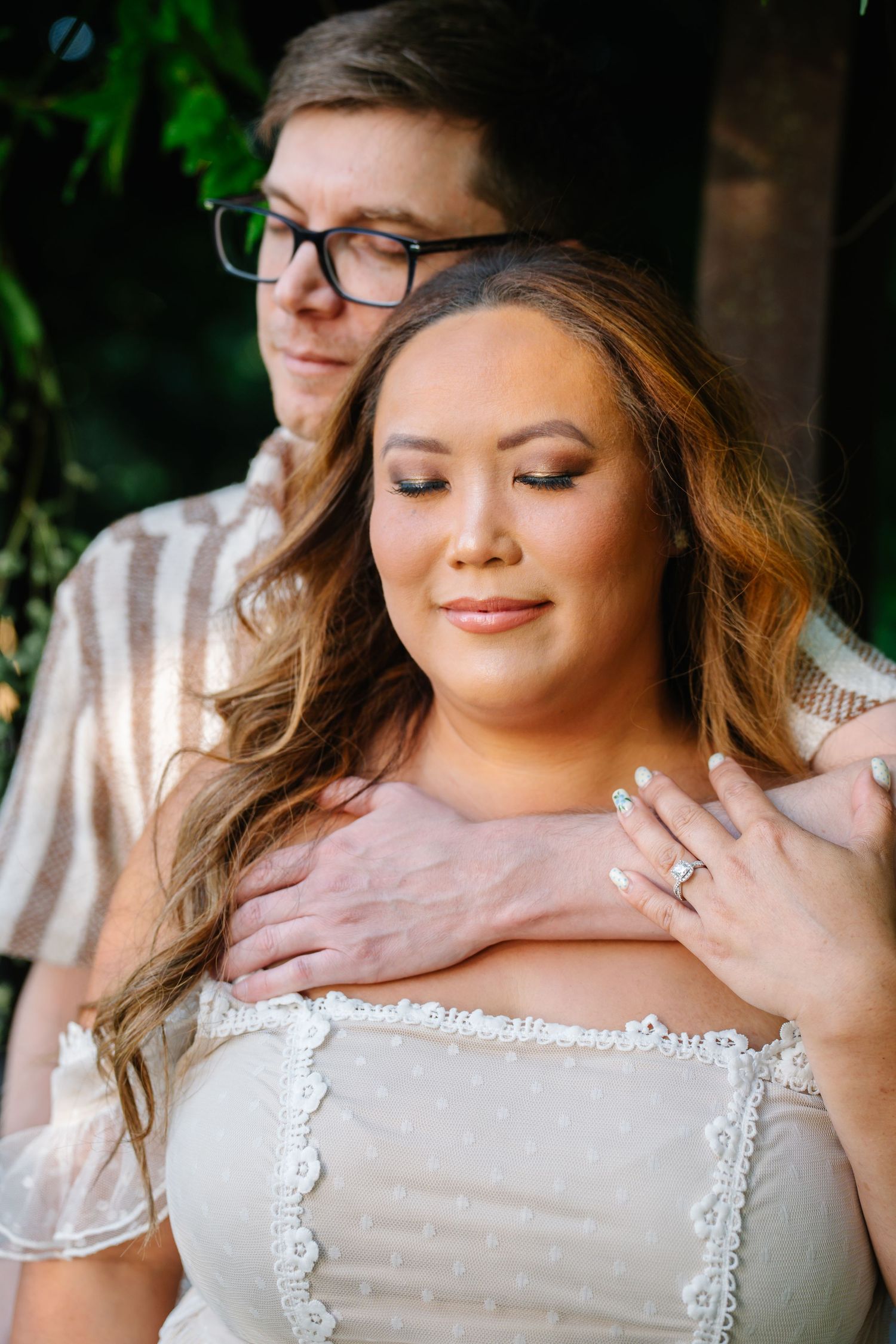 Couple in romantic pose, woman in white lace dress with engagement ring, man in striped shirt with glasses outdoors.