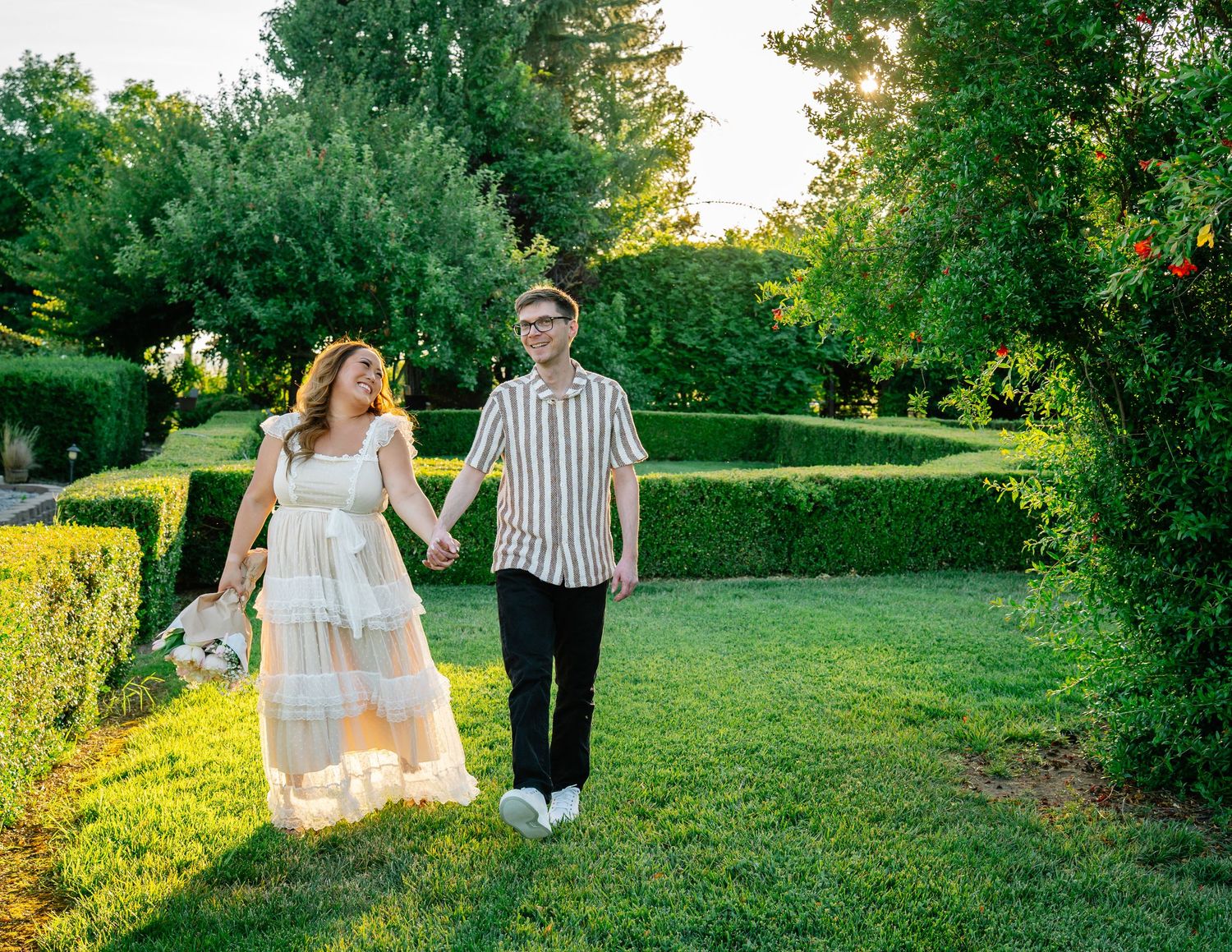 Couple holding hands walking through lush green garden at golden hour, woman in white dress, man in striped shirt.