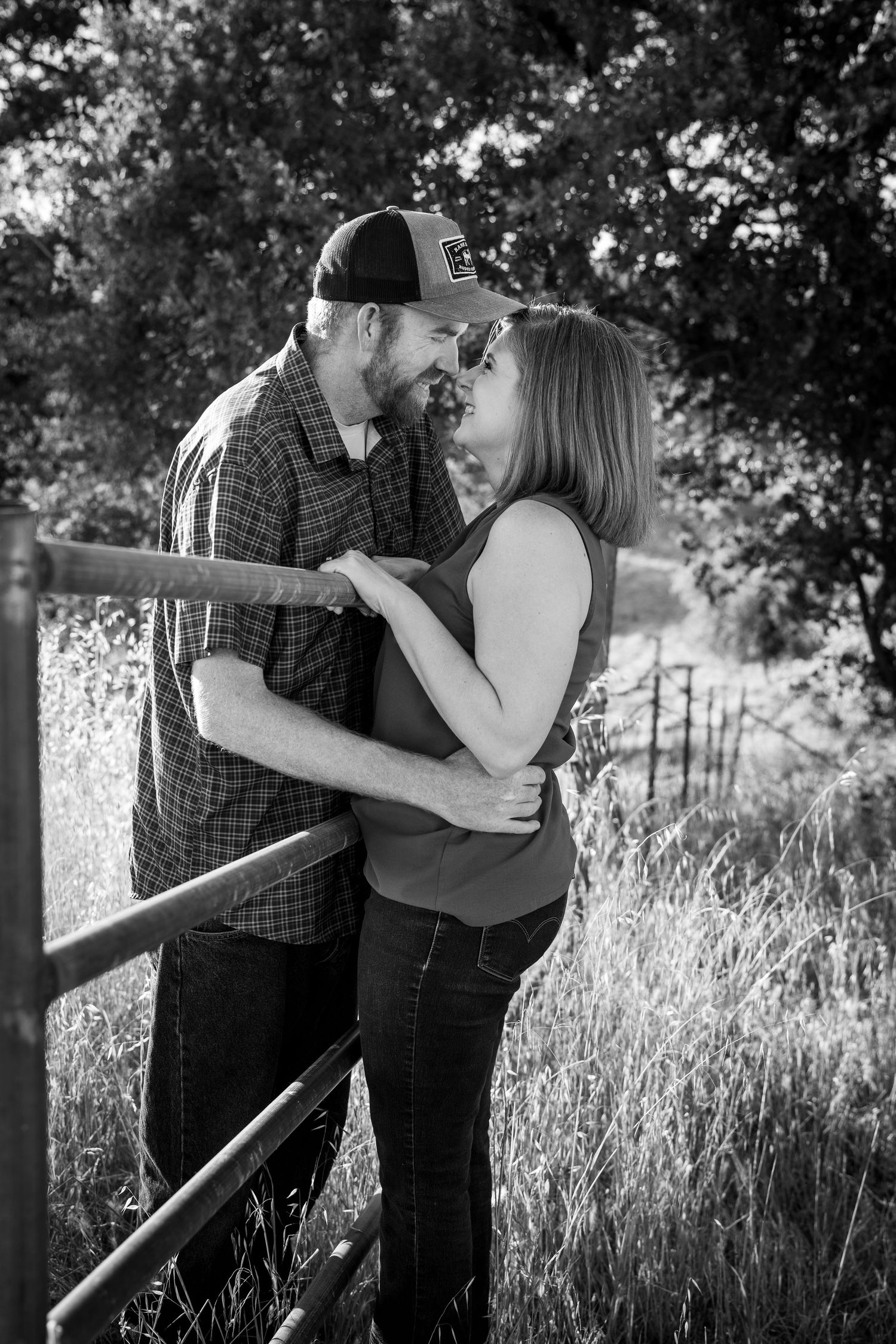 Black and white photo of a couple embracing and about to kiss near a rustic fence surrounded by tall grass and trees.