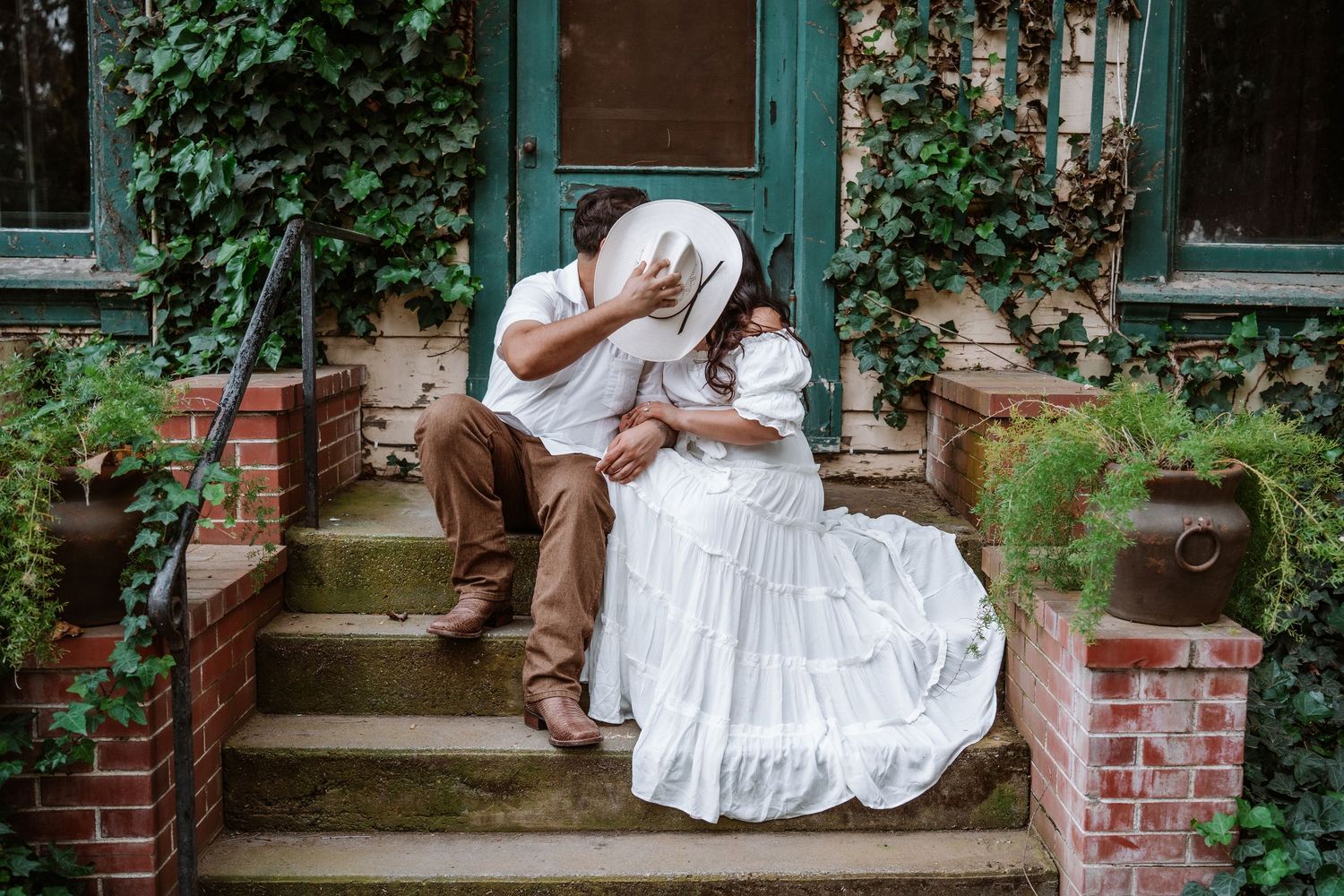 Couple sharing a romantic kiss on brick steps outside a ivy-covered home, woman in white dress.