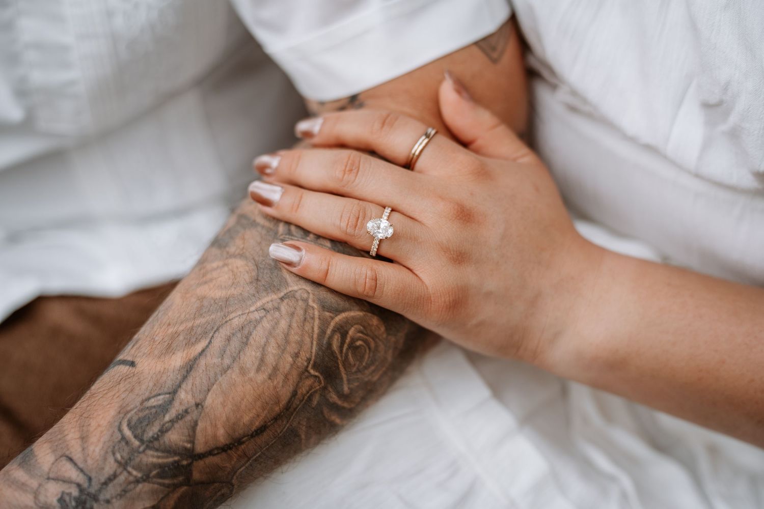 Close-up of a woman's hands wearing a diamond engagement ring and gold band, with a floral tattoo visible on her arm.