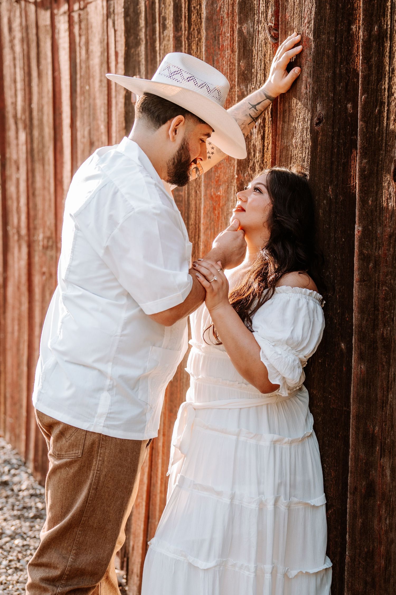 Romantic couple in white outfits leaning against rustic wooden wall, man in cowboy hat holding woman's hand.