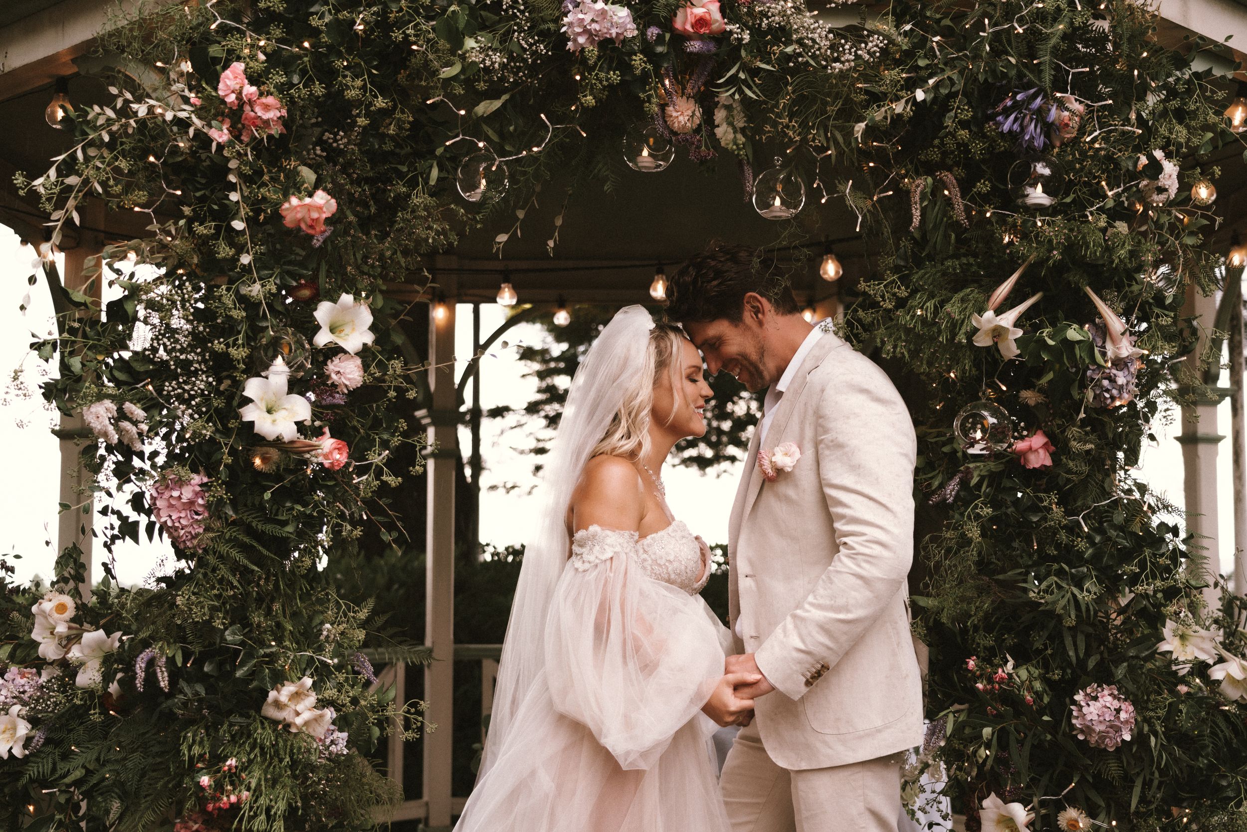 Couple embraces under floral arch during romantic outdoor wedding ceremony.