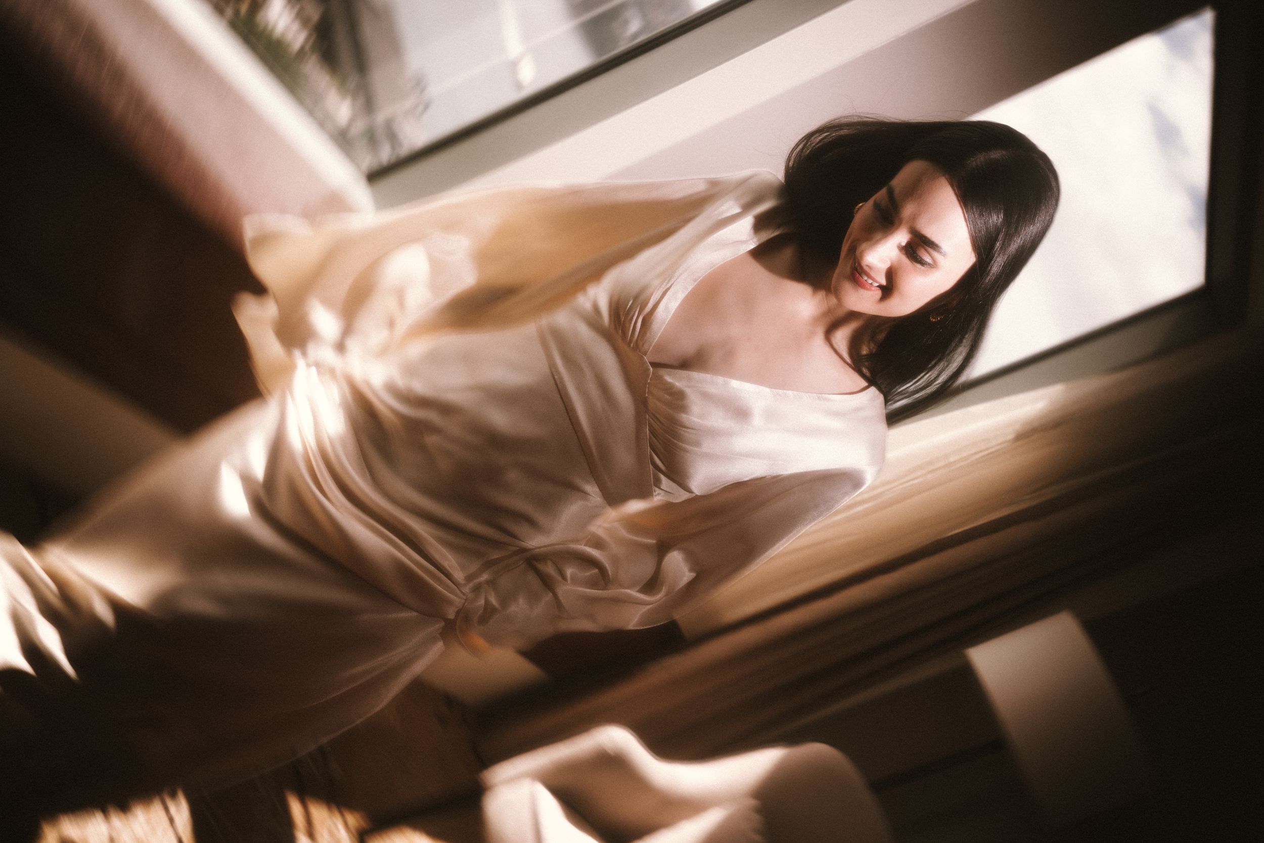 Bride in elegant silk dress illuminated by dramatic window light.