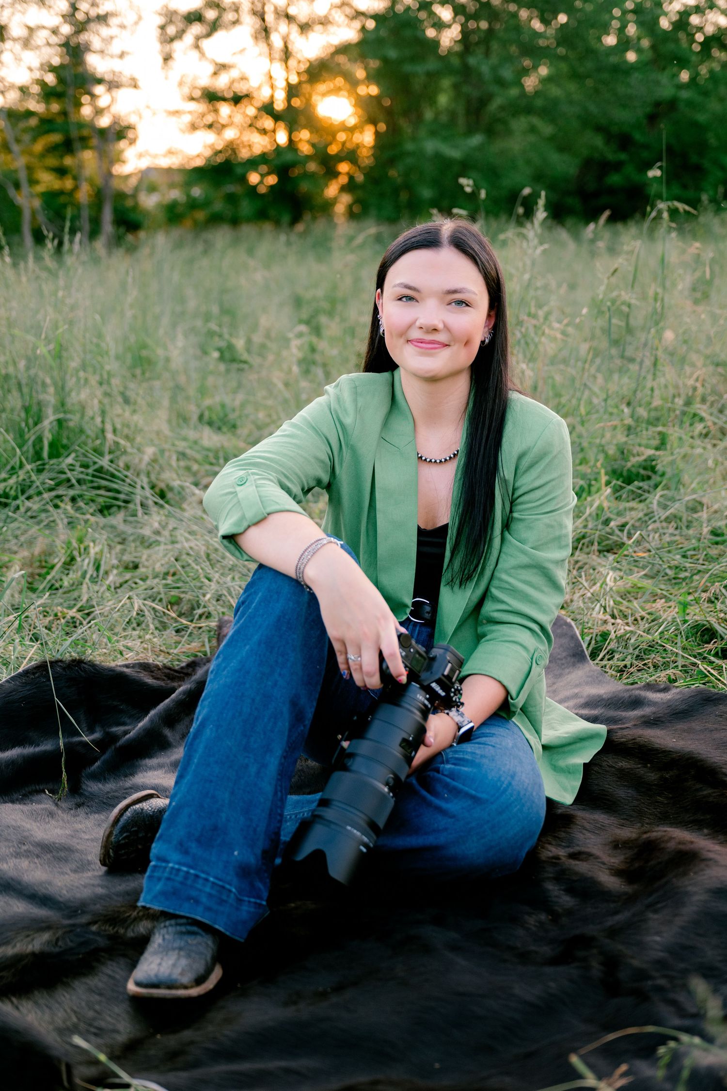 Smiling female photographer holding a camera and tripod outdoors in a lush green field at golden hour sunset.