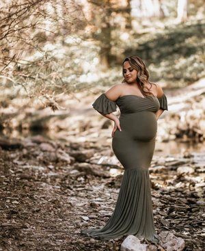 Maternity photo shows pregnant woman in olive off-shoulder gown posing gracefully in a sunlit wooded setting. At a photoshoot in Bexley