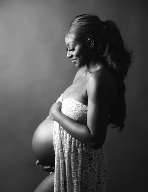 Black and white maternity portrait showing a pregnant figure in lace dress with flowing hair against dark backdrop.