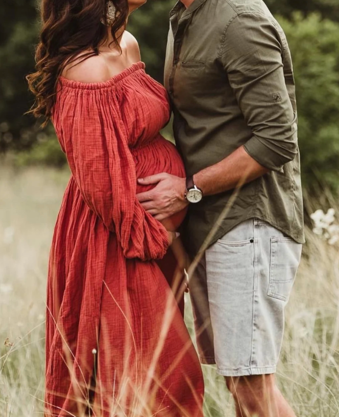 A couple stands together in a sunny field, her wearing a flowey red dress and him in a green button-up shirt and shorts in Bexley