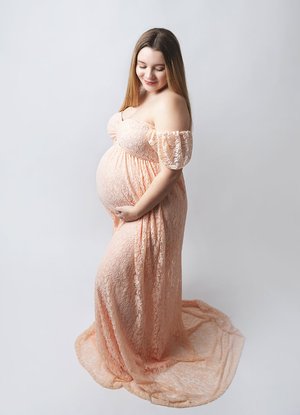 Elegant maternity portrait featuring an off-shoulder peach lace gown against a light grey backdrop. During a maternity session in Bexley