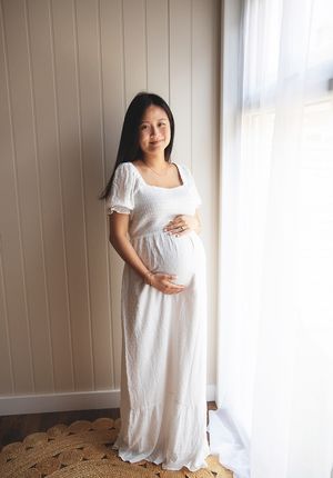 An expectant woman in a flowing white maxi dress stands by a window in soft natural light. During a maternity photoshoot in London