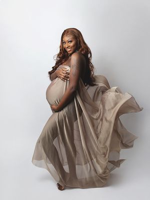 Side view of an expectant parent in a flowing taupe sheer dress gracefully posing against a white backdrop during a maternity shoot.