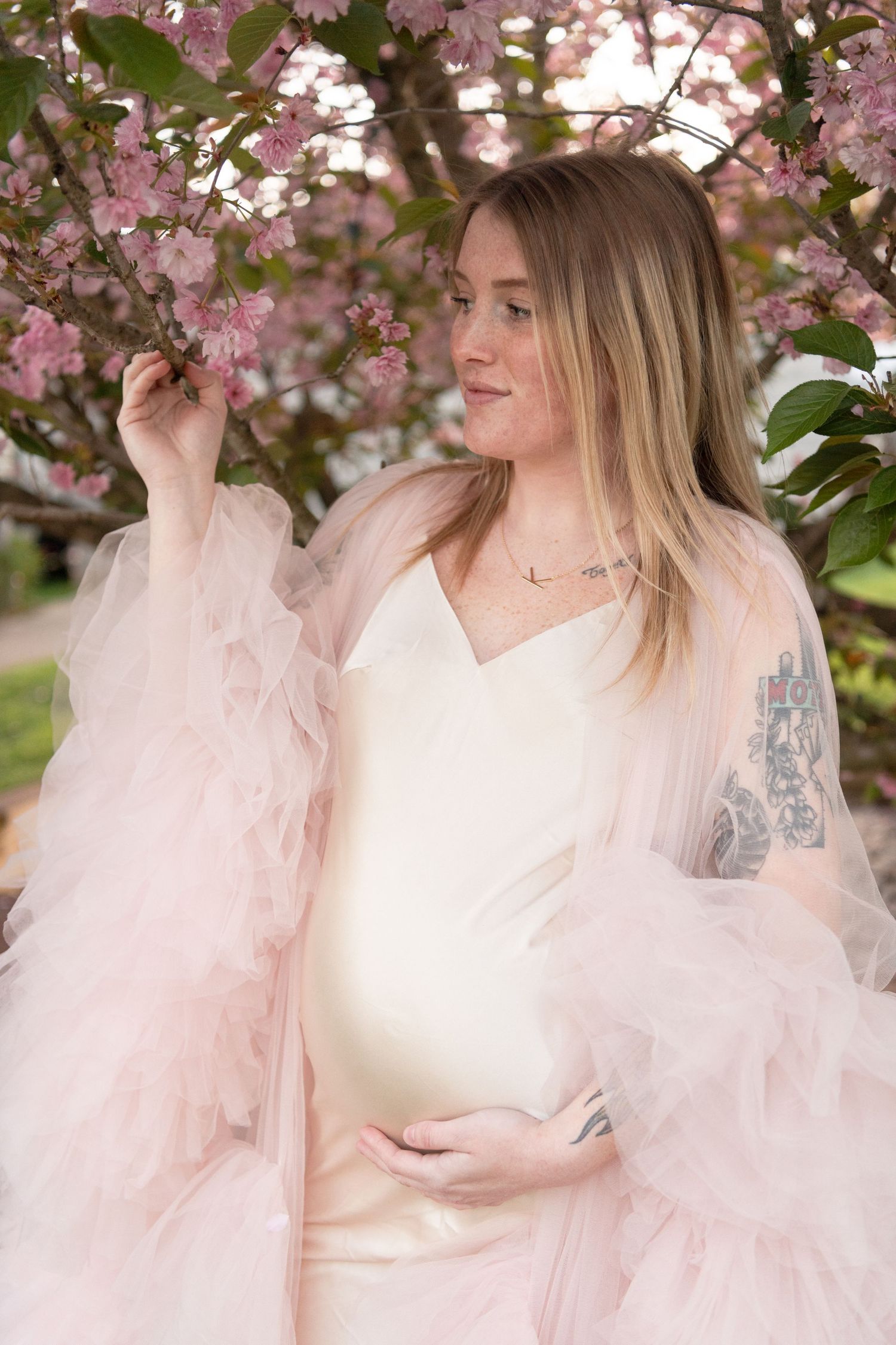 Expectant mother in flowing pink dress poses among cherry blossoms in spring.