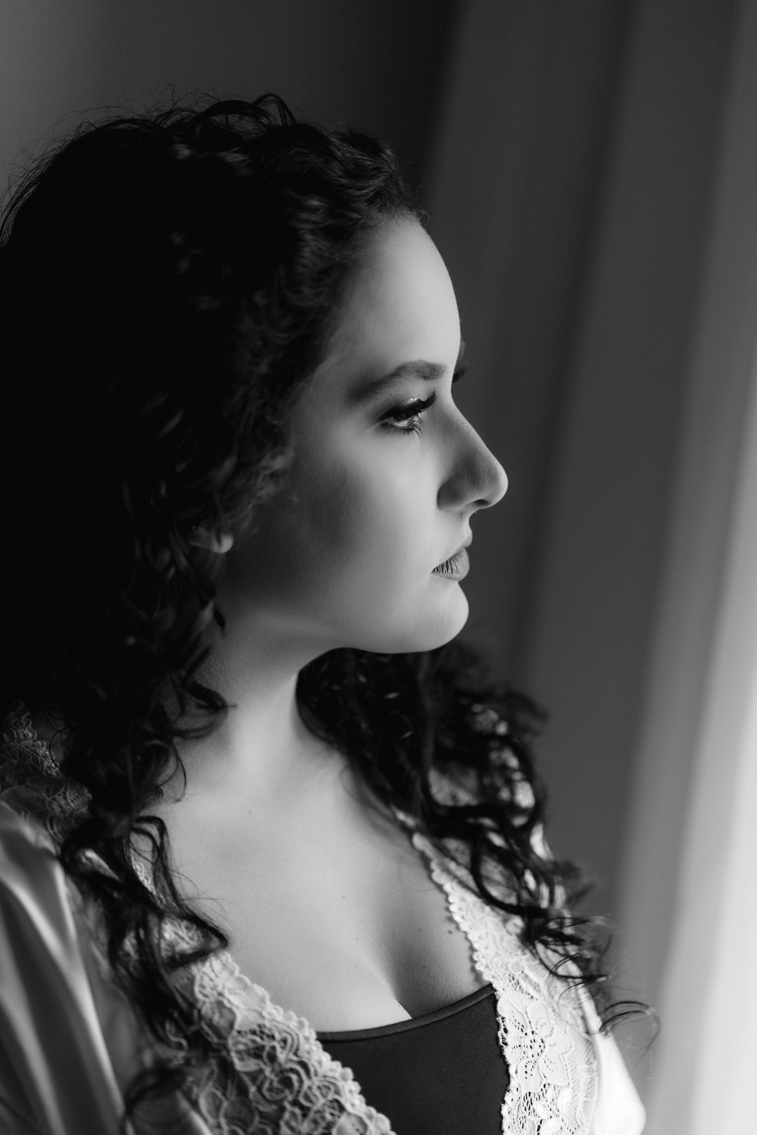 Black and white profile portrait of woman with curly hair in lace garment near window.