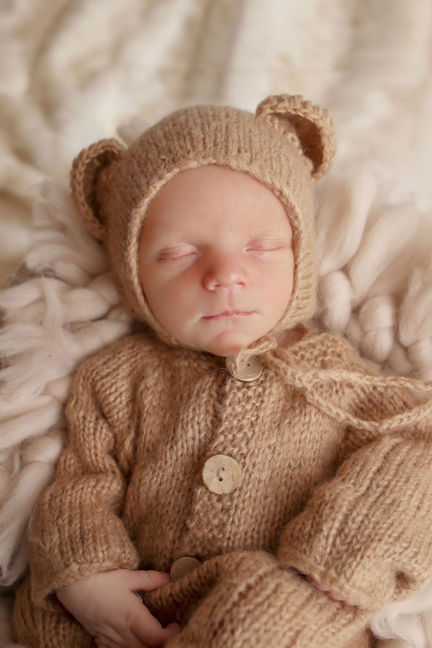 Sleeping newborn wearing cozy knitted brown bear outfit with ears on cream blanket.
