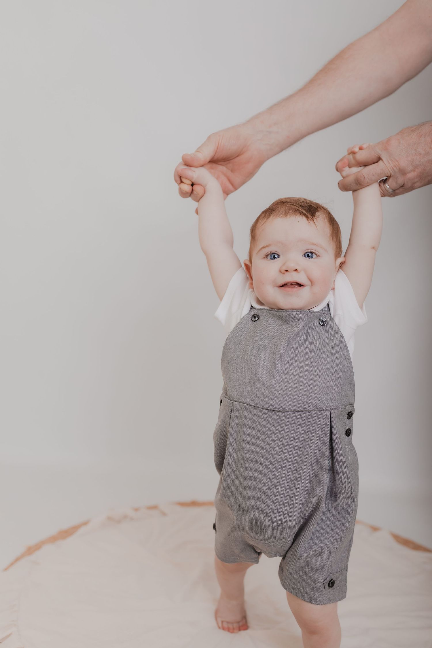 Baby wearing grey romper outfit being held up by adult hands against white backdrop. Photographer. Hull. East Yorkshire. Photography