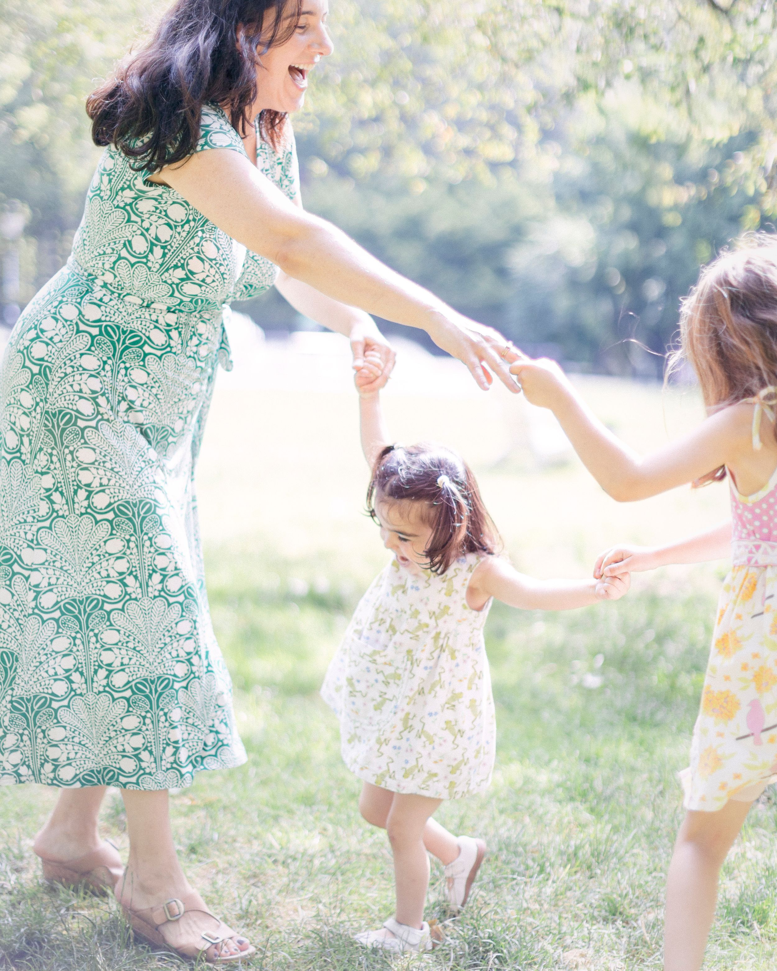 A family plays together outdoors in summer dresses, dancing and holding hands in sunlit grass.