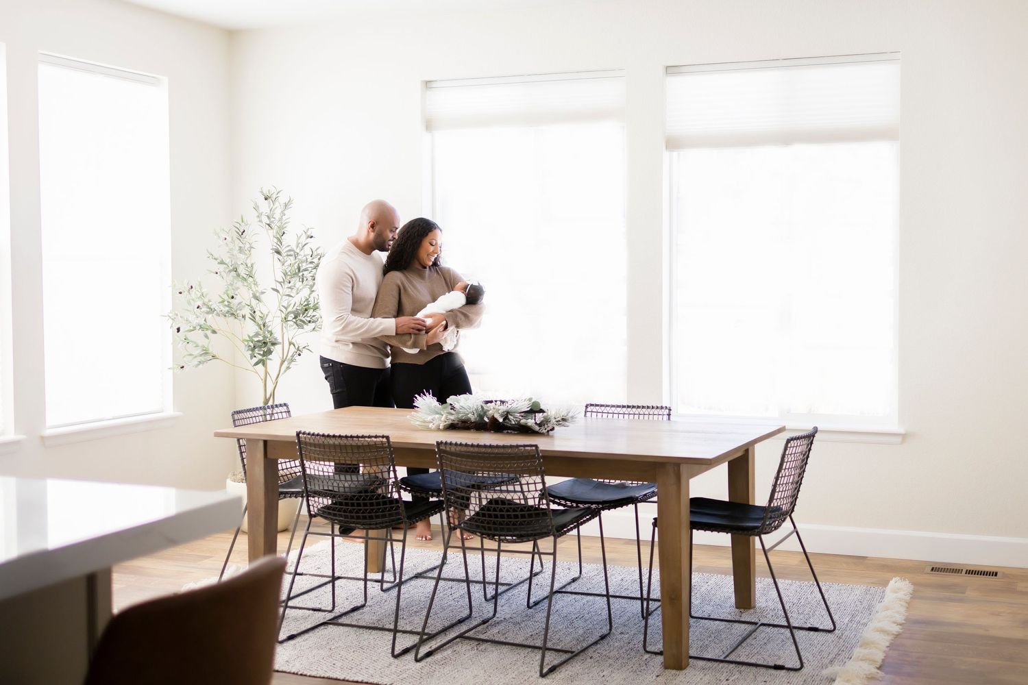 Man and woman stood in their dining room, next to bright windows, woman holding newborn girl,both looking at her smiling