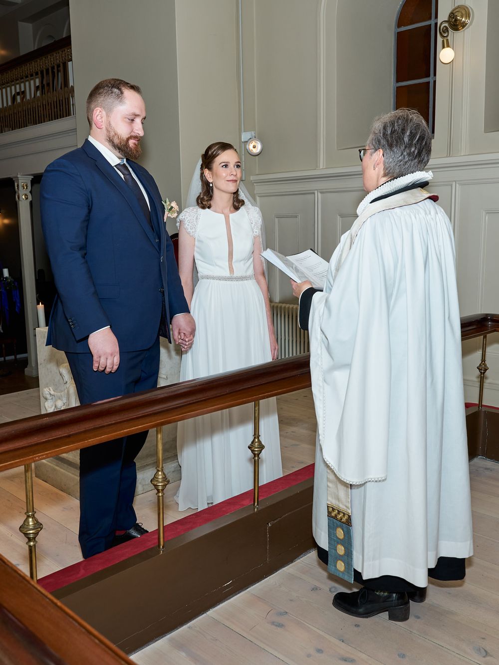 Wedding ceremony with couple in white dress and navy suit being officiated by priest in church.