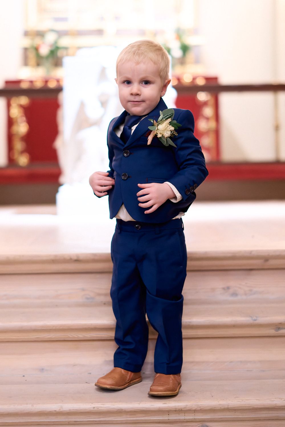 Toddler in navy blue suit with boutonniere stands in church setting.