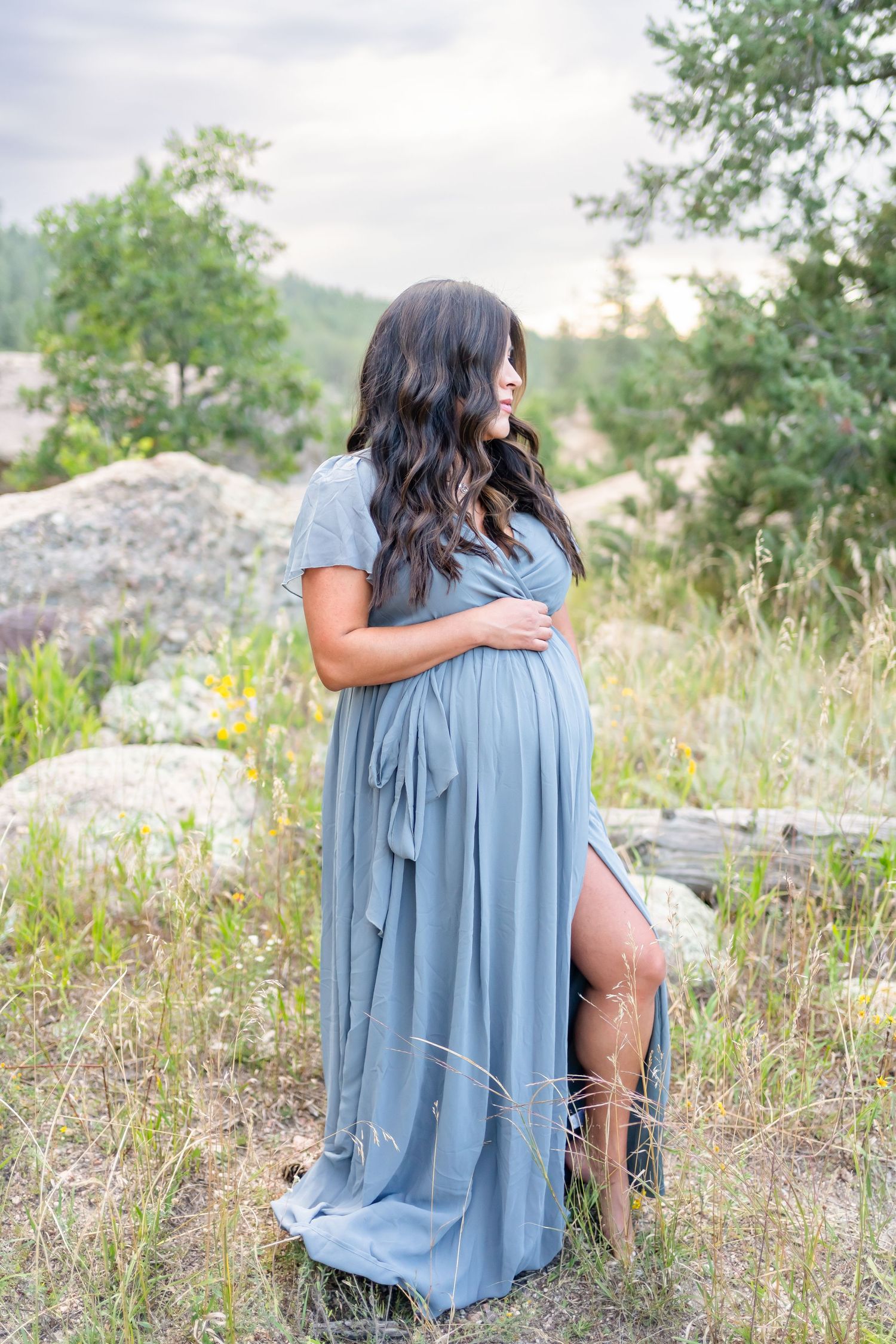 Maternity photo in blue flowing dress posed against natural rocky outdoor setting at sunset.