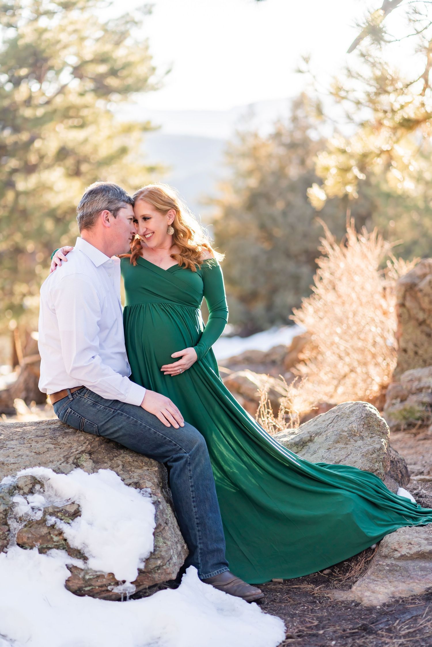 Romantic winter maternity shoot with couple on snowy rocks wearing green dress and white shirt.