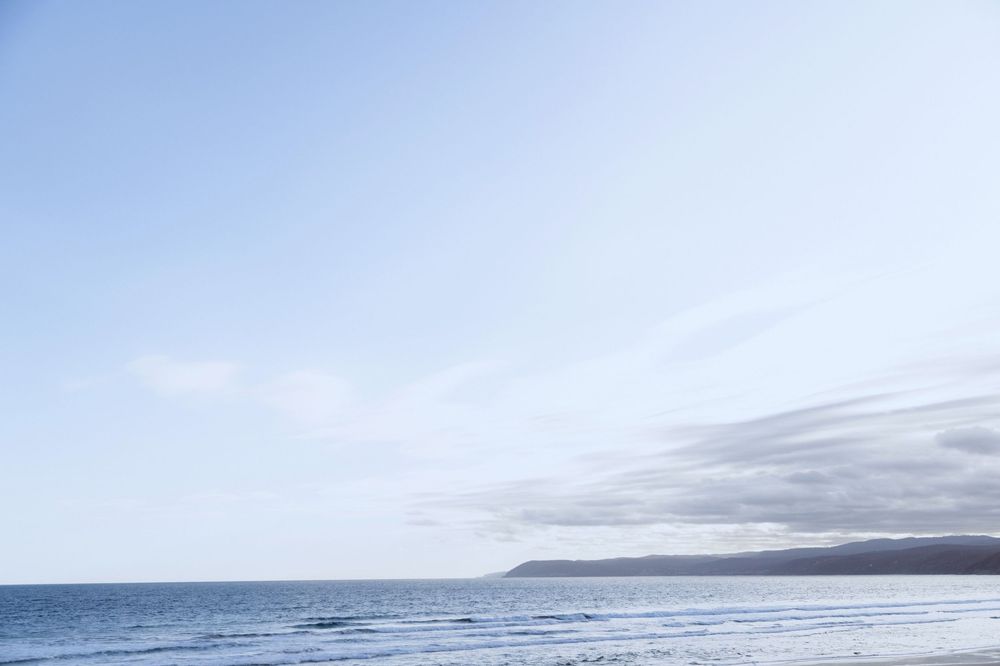 A scenic panoramic view of waves crashing on a rocky beach with mountains and cloudy skies in the distance.