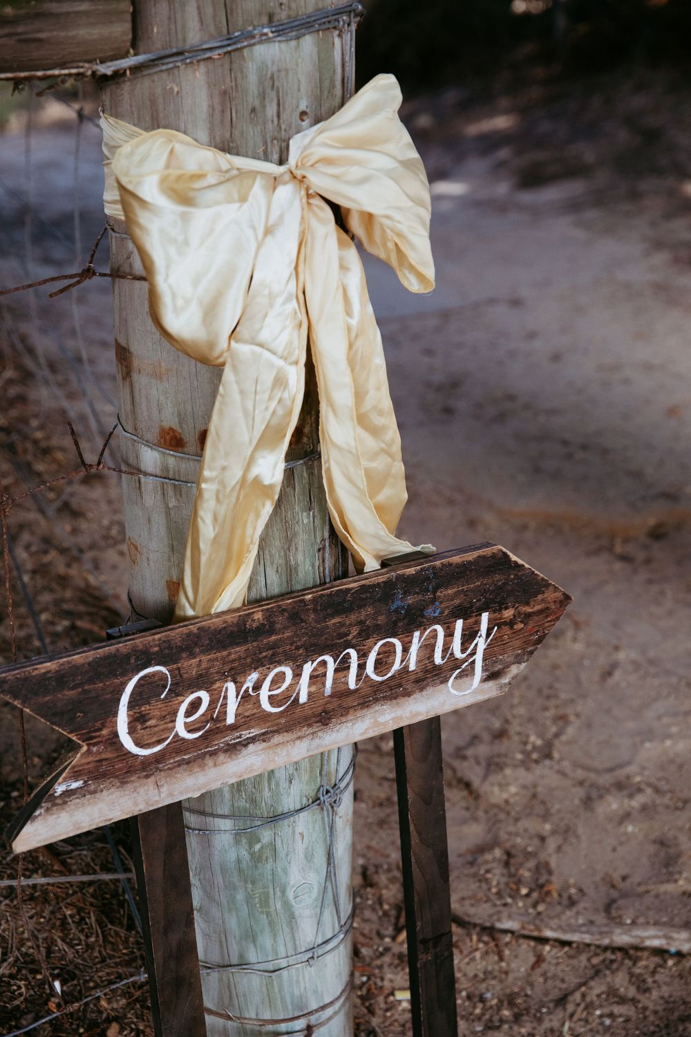 Rustic wooden ceremony sign decorated with cream-colored fabric bow for outdoor wedding decor.