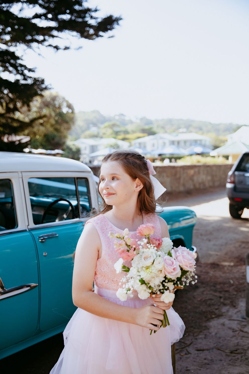 Someone in a pink dress holds a delicate bouquet of flowers next to a vintage turquoise car on a sunny day.