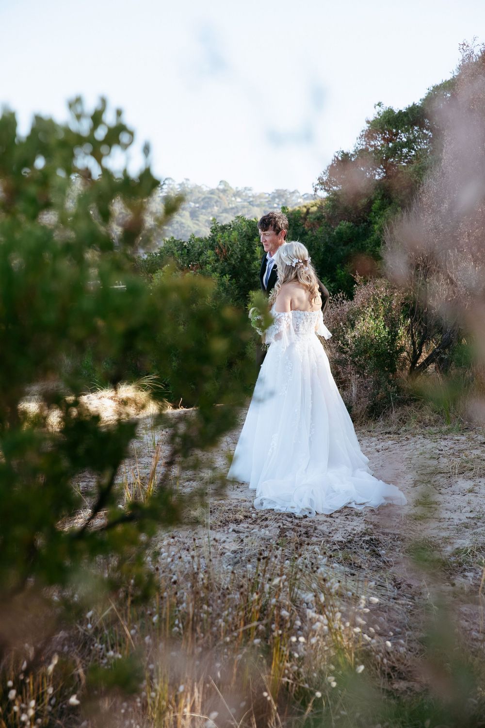 A bride in a flowing white wedding dress walks through a rustic outdoor setting with wildflowers and natural foliage.