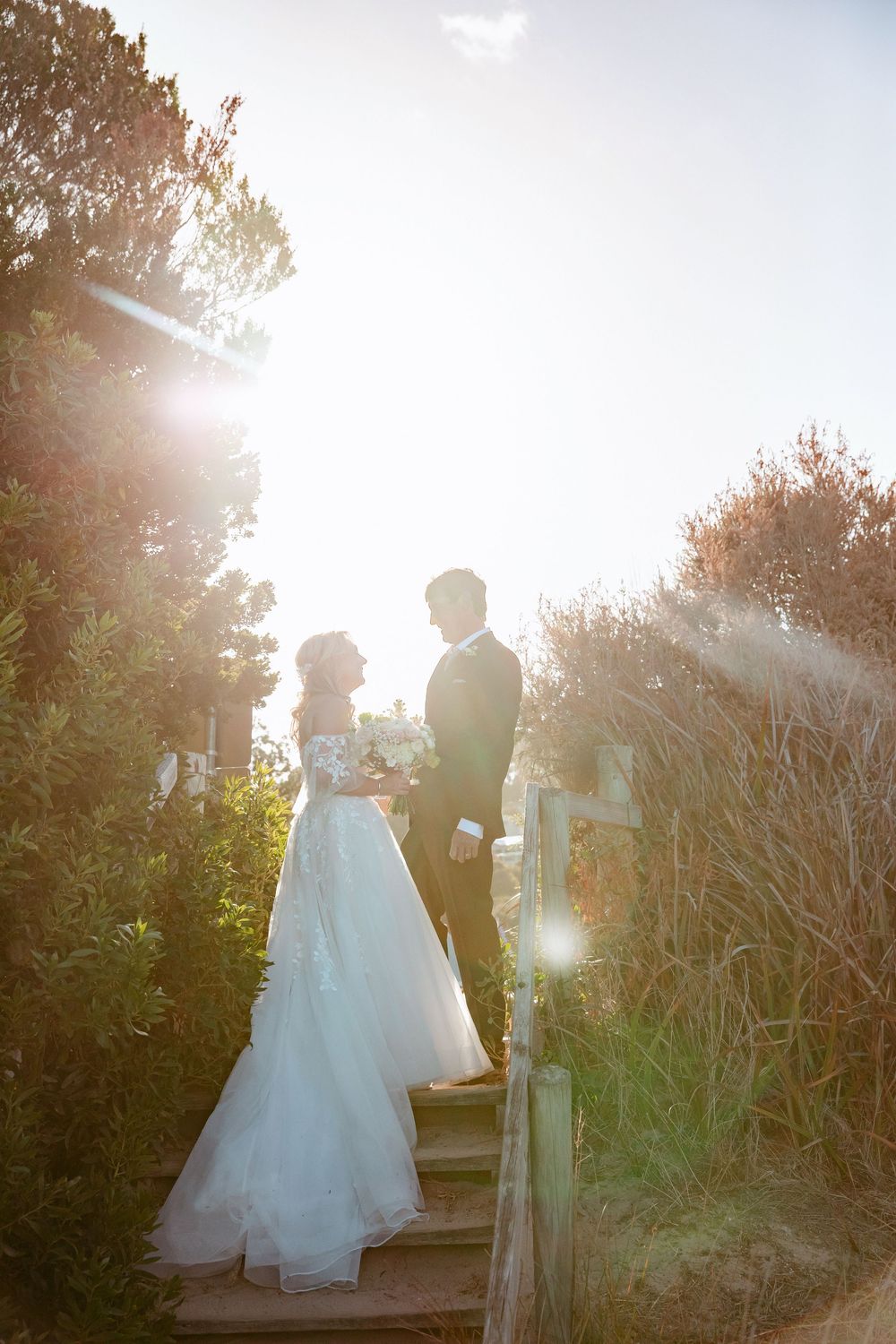 A romantic wedding couple stands in golden sunlight on a garden path during sunset, creating a dreamy silhouette effect.