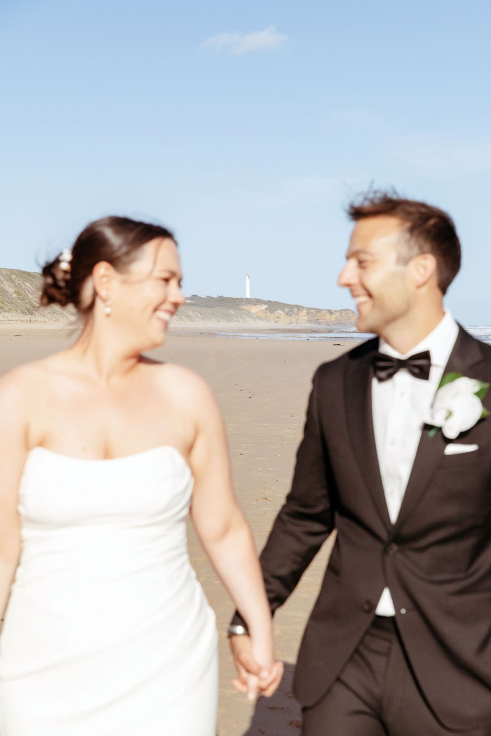 A happy couple in wedding attire walks and laughs together on a beach during their special day.