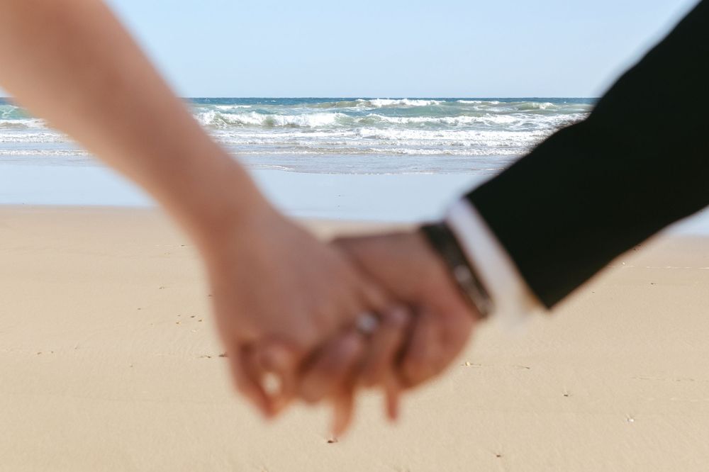 A couple holds hands while walking on a sandy beach with ocean waves in the background.
