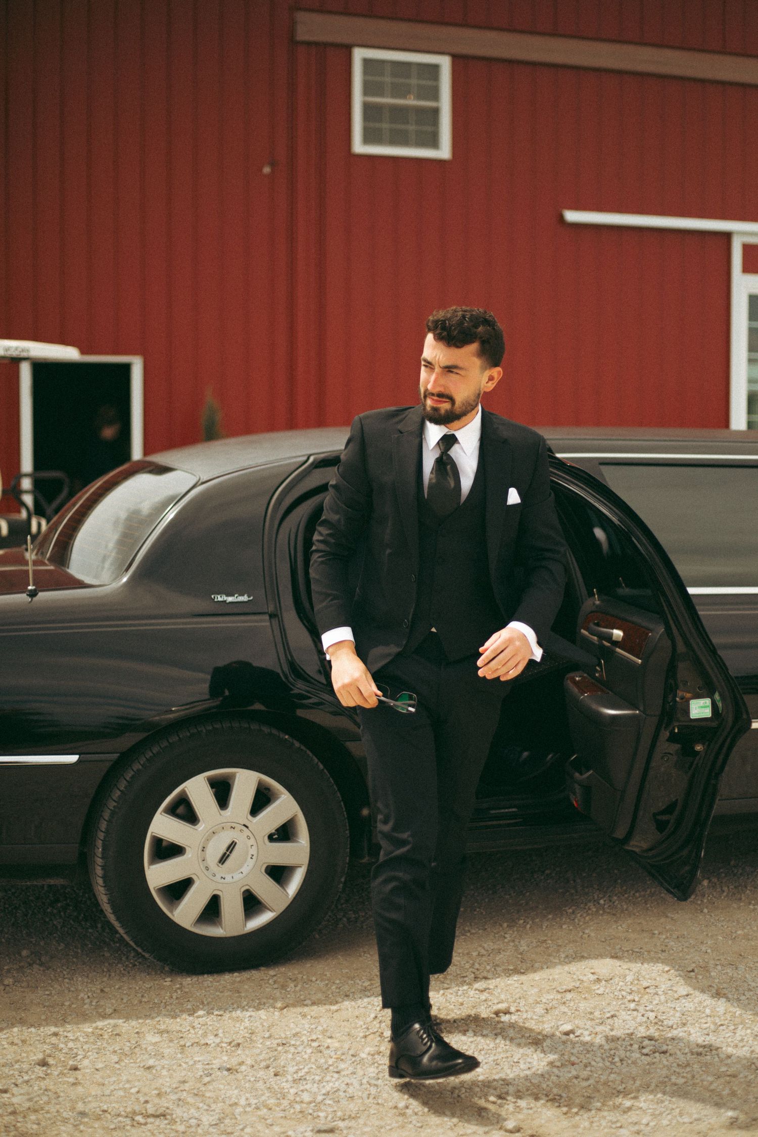 Stylishly dressed man in black suit exits luxury car near red barn building.